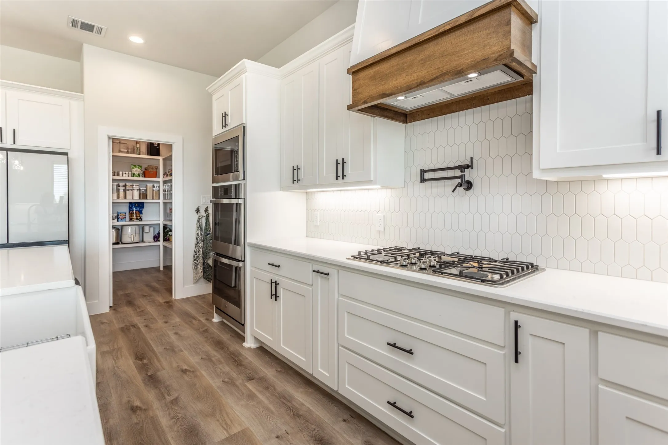 White cabinetry with black hardware, wood-finish flooring, white countertops, built-in double ovens, and a gas cooktop with a pot filler