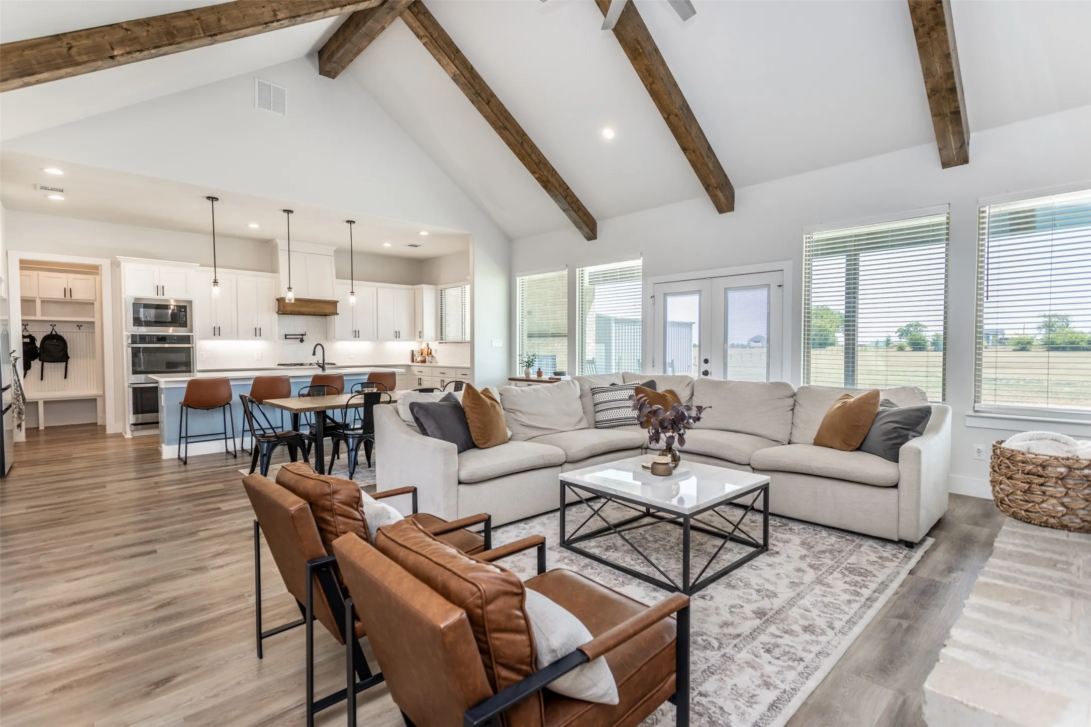 Vaulted ceiling with exposed wood beams, wood-finish flooring, white cabinetry with integrated appliances, kitchen island with seating, and multiple windows