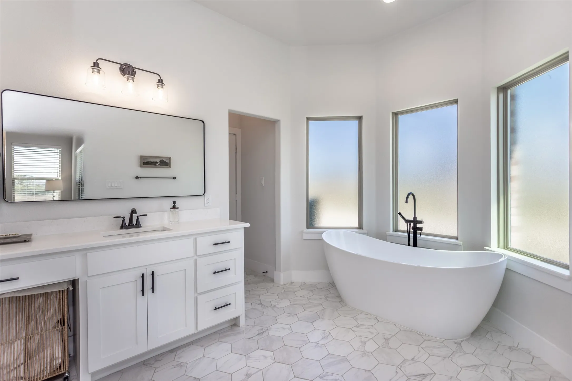 Freestanding soaking tub, white vanity with integrated sink, stone-finish hexagonal floor tiles, and multiple windows with frosted glass