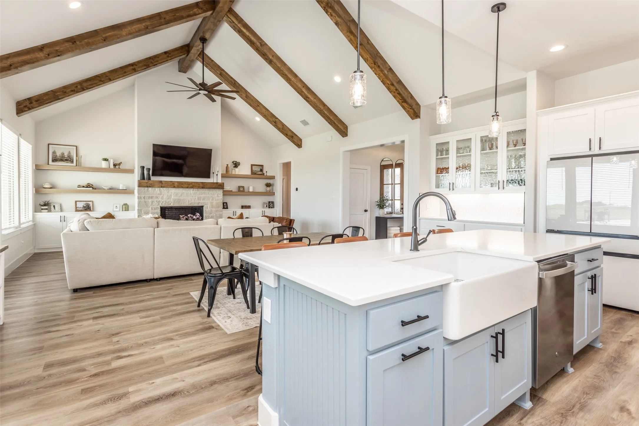 Vaulted ceiling with exposed wood beams, wood-finish flooring, white stone fireplace, and a kitchen island with a farmhouse sink and stainless steel appliances