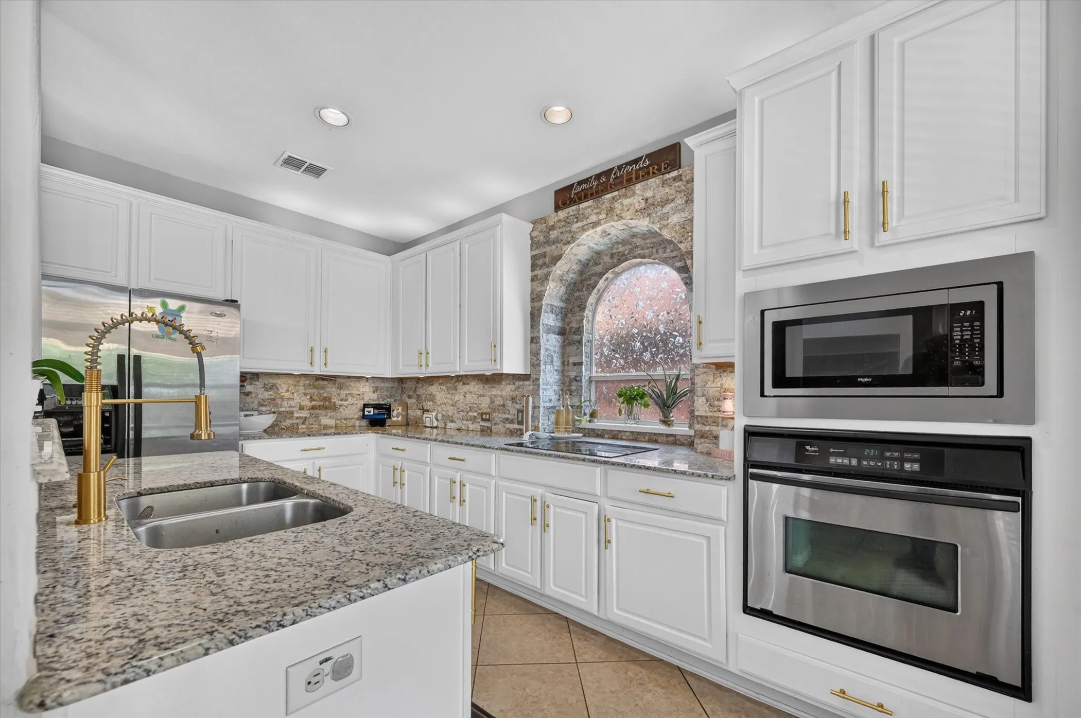 White cabinetry with gold-finish hardware, granite countertops, stainless steel appliances, dual basin sink with gooseneck faucet, and a stone-finish arched window surround