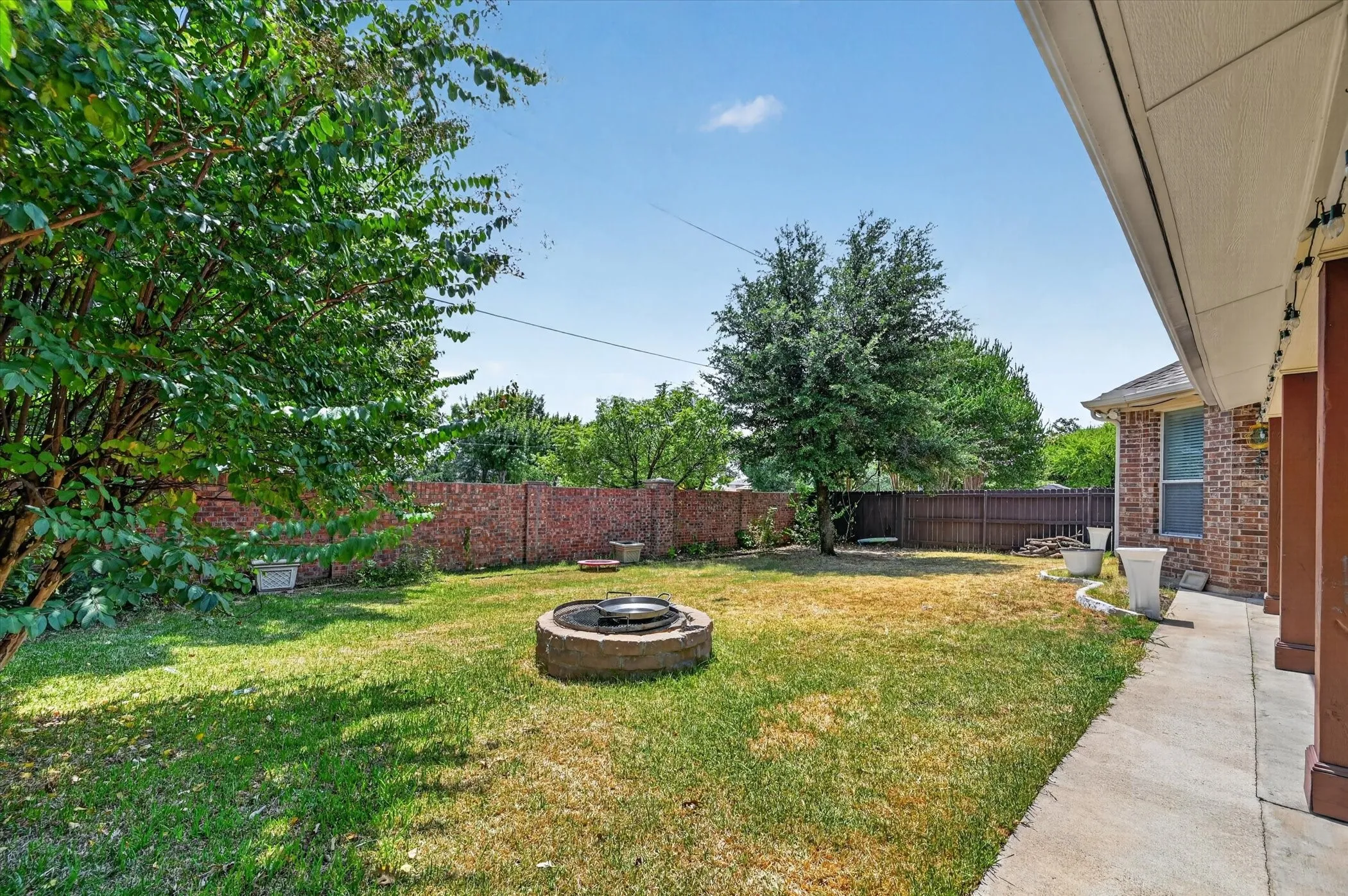 Private backyard featuring a brick privacy fence, mature trees, concrete patio, and a built-in fire pit