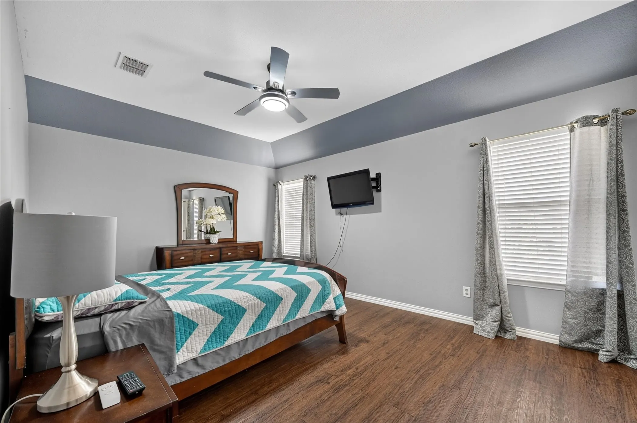 Tray ceiling with accent paint, wood-finish flooring, two windows with blinds, and a ceiling fan with integrated lighting