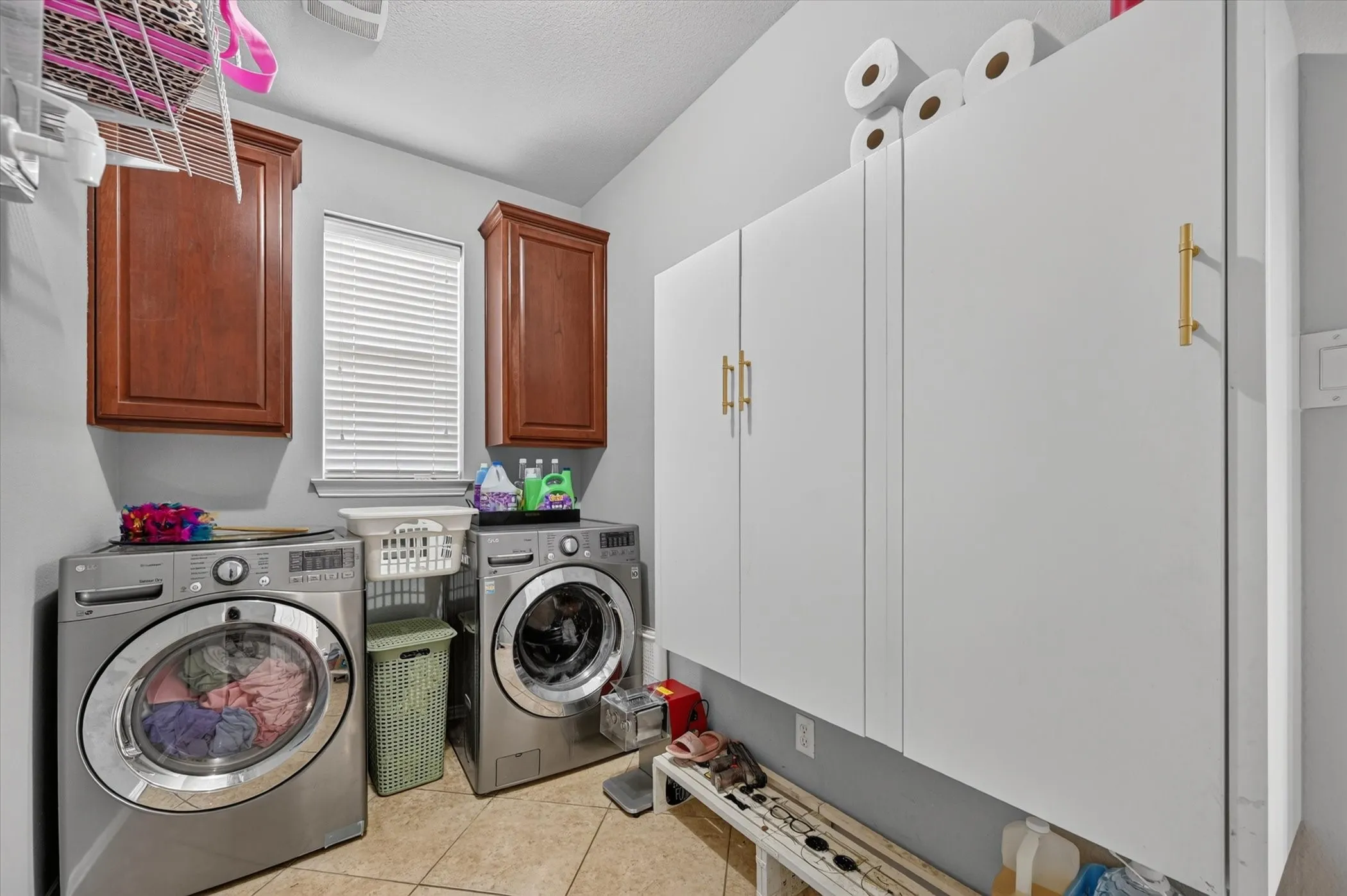 Laundry room featuring tile flooring, a window with blinds, and white shaker-style cabinetry with gold-tone hardware