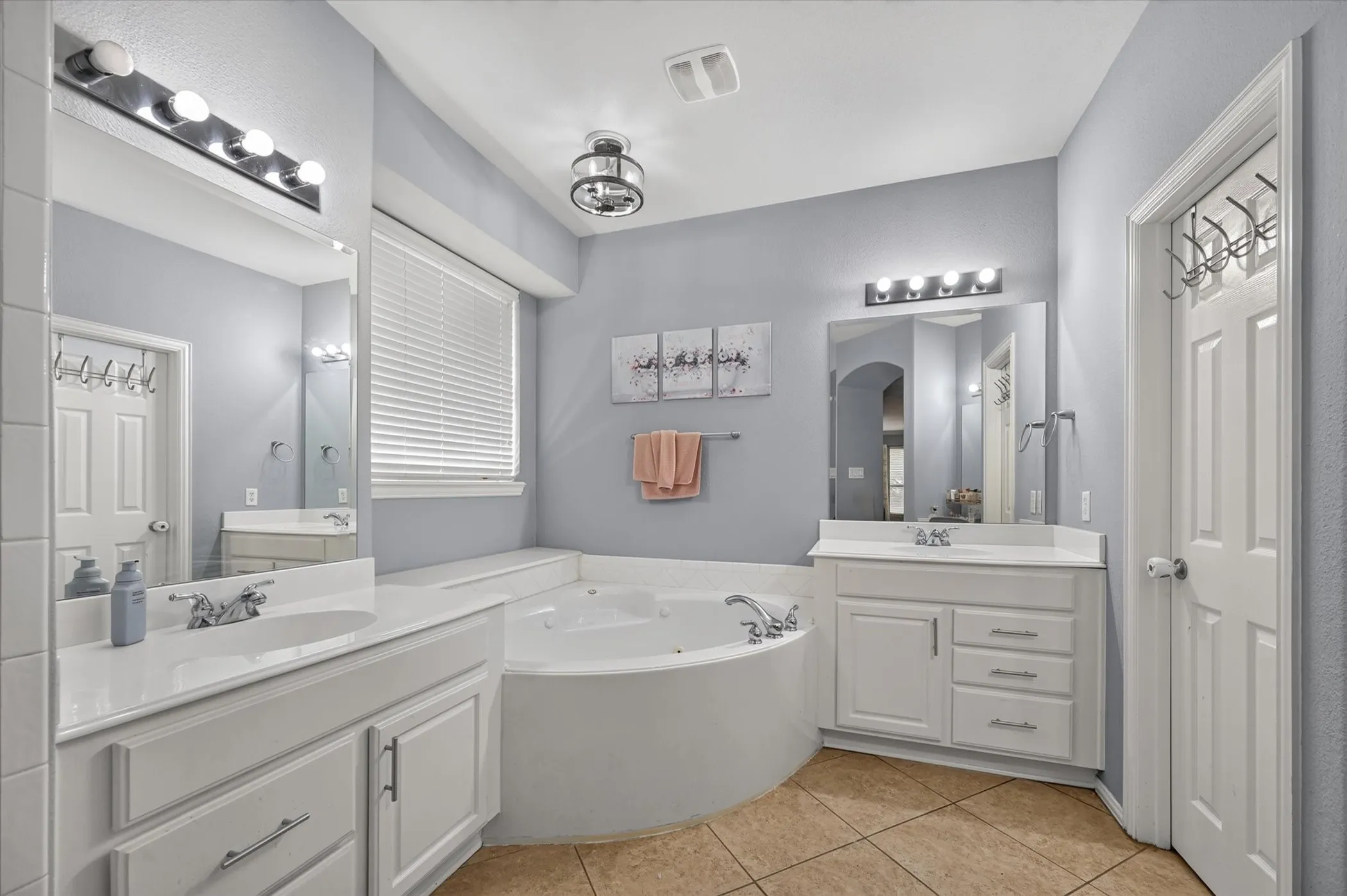 Two white vanities with integrated sinks, a built-in jetted tub, tiled flooring, a window with blinds, and flush-mount lighting fixtures