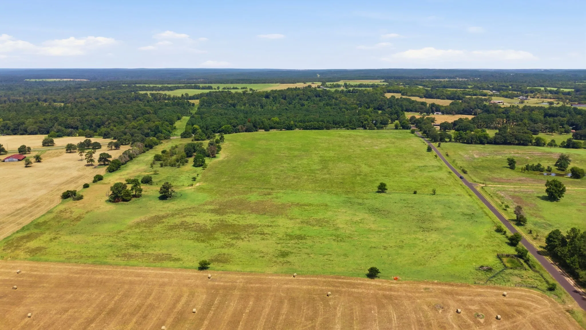 Expansive rural landscape featuring open fields, distant wooded areas, and a paved road