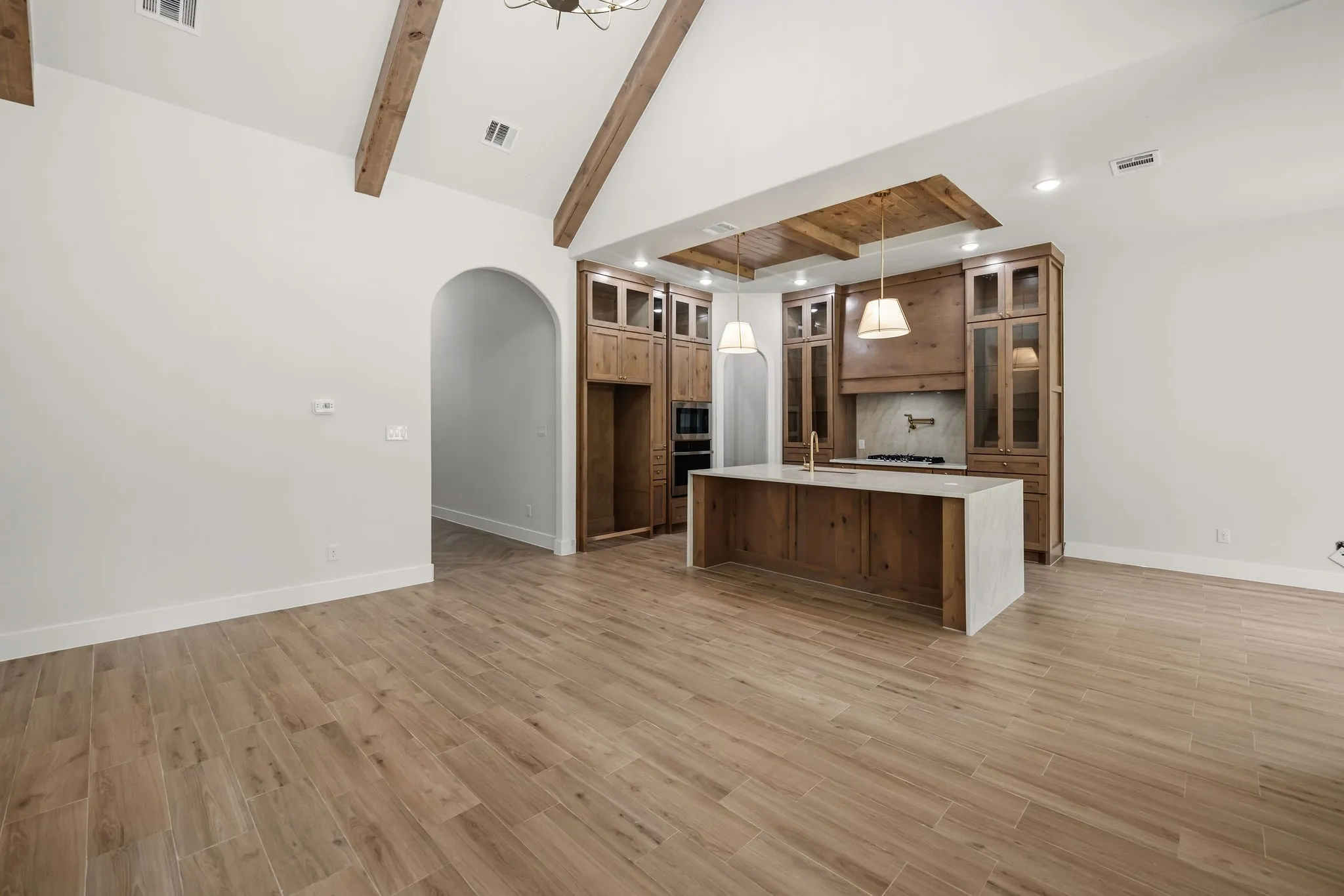 Vaulted ceiling with exposed wood beams, wood-finish flooring, custom wood cabinetry, kitchen island with a stone countertop, and arched entryway