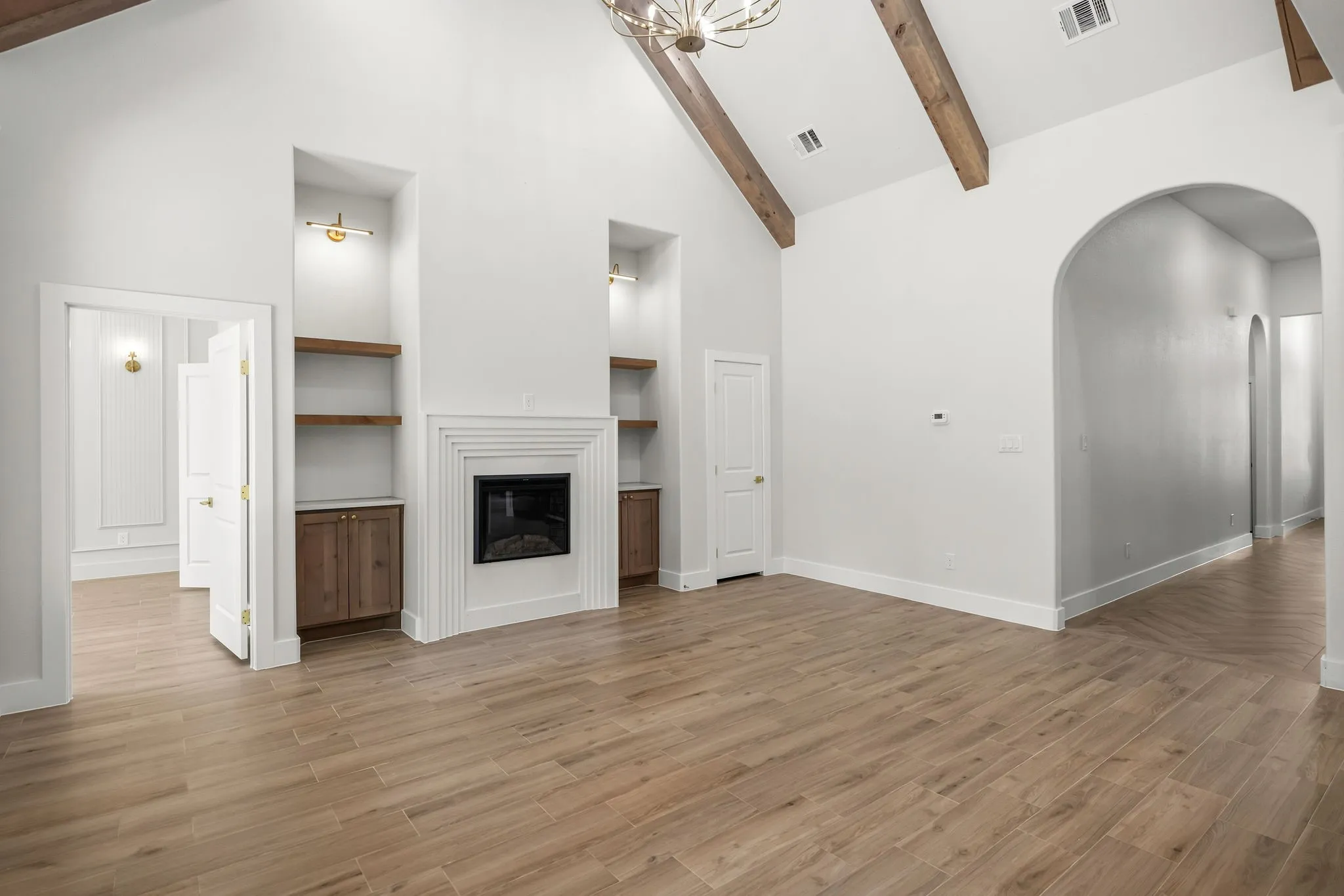 Vaulted ceiling with exposed wood beams, wood-finish flooring, built-in shelving with integrated lighting, fireplace, and arched doorways