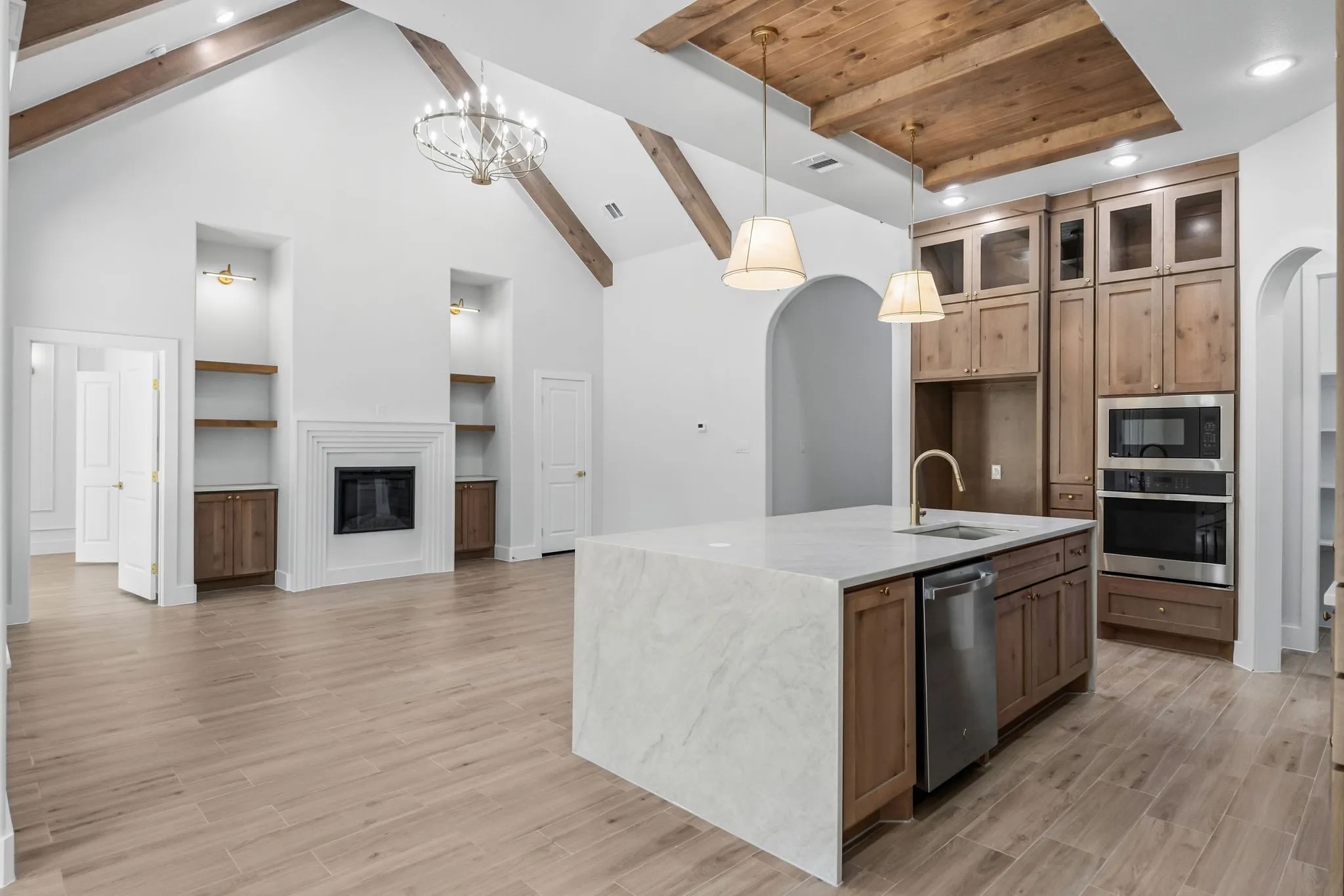 Vaulted ceiling with exposed wood beams, central fireplace flanked by built-in shelving, kitchen island with stone finish countertop, built-in stainless steel appliances, and wood-finish cabinetry