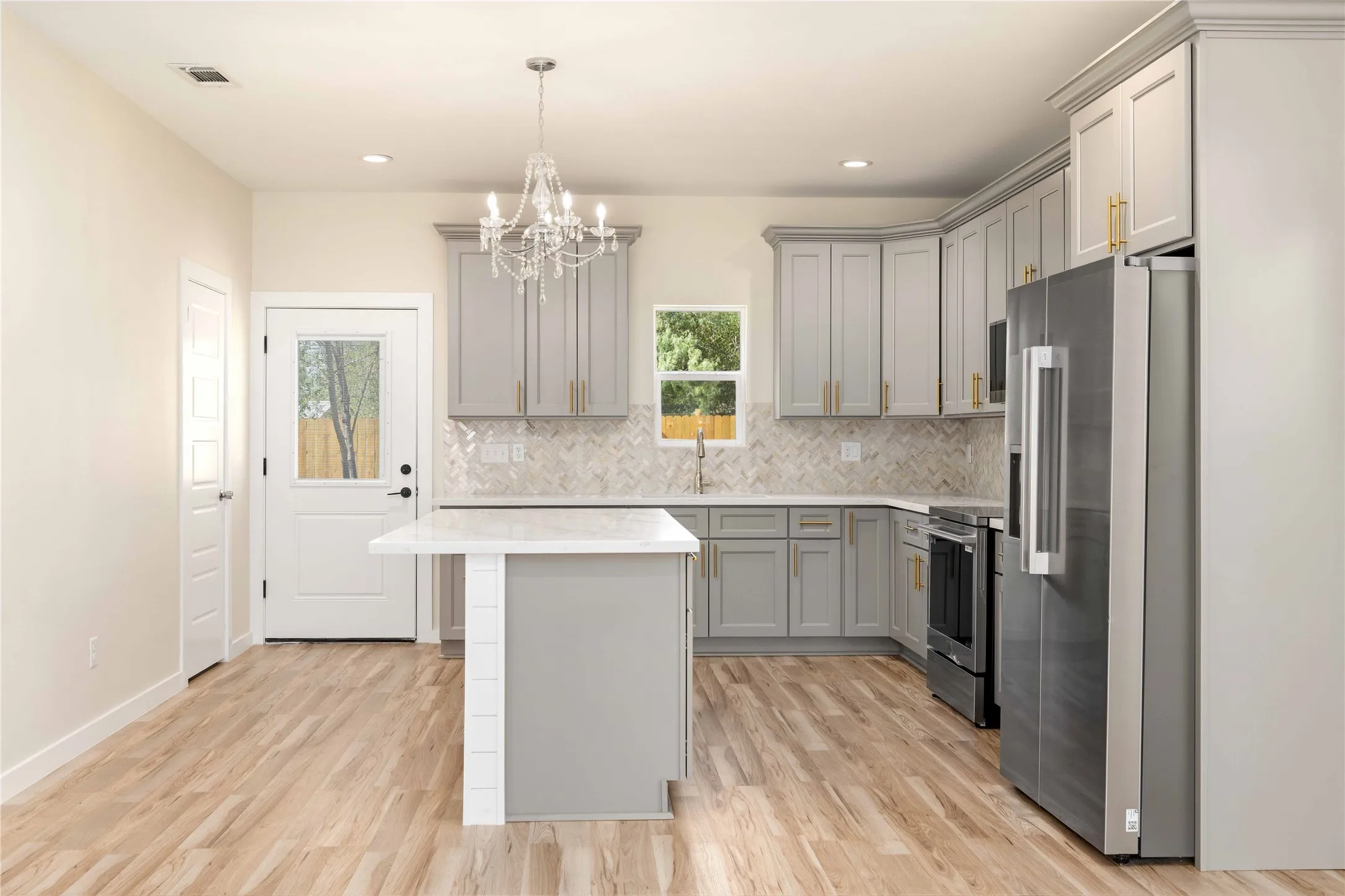 Kitchen featuring wood-finish flooring, light grey cabinetry with gold-toned hardware, a central island with a white countertop, stainless steel appliances
