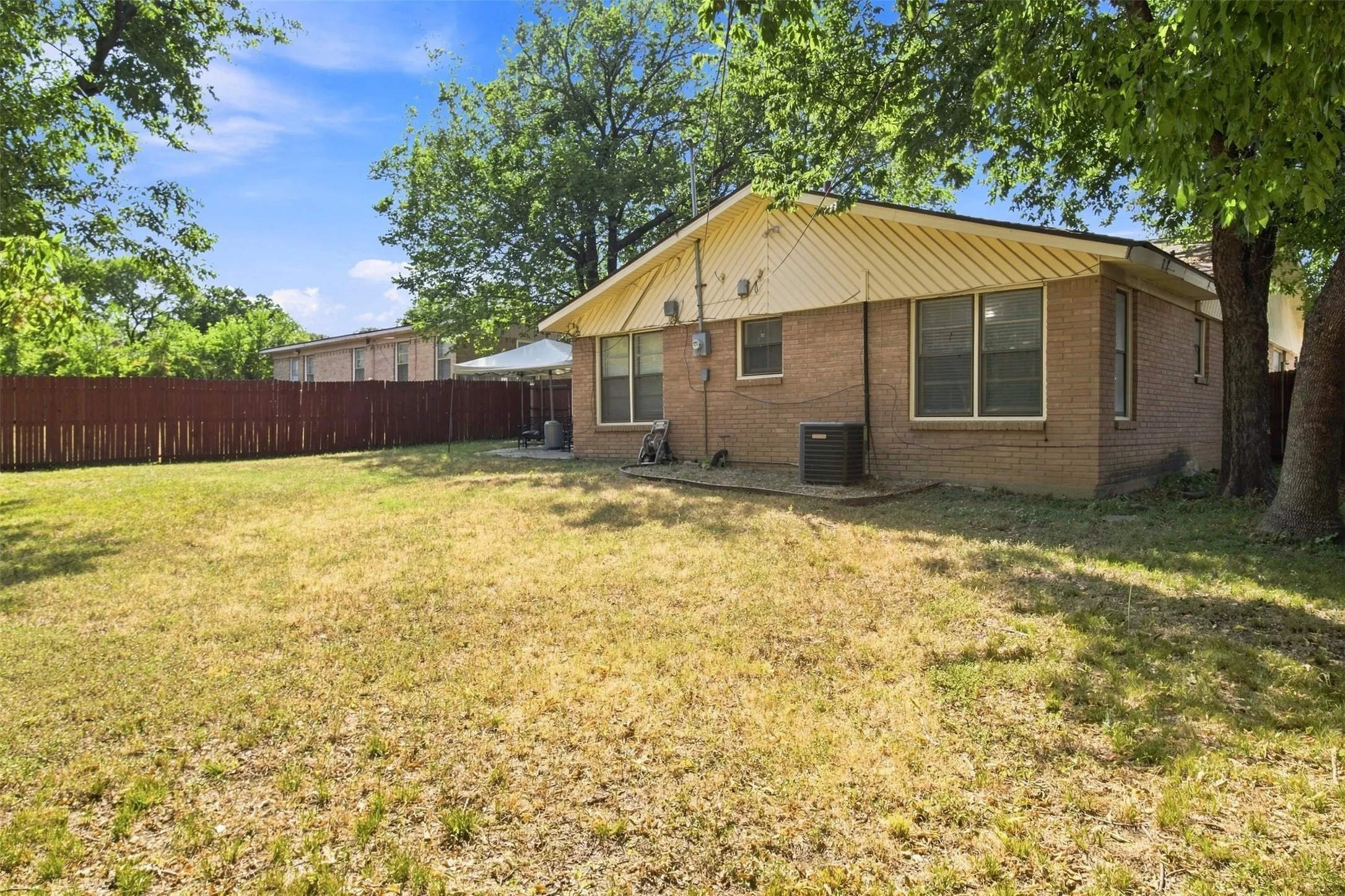 Expansive grassy yard with a privacy fence, brick exterior, multiple windows, and mature trees
