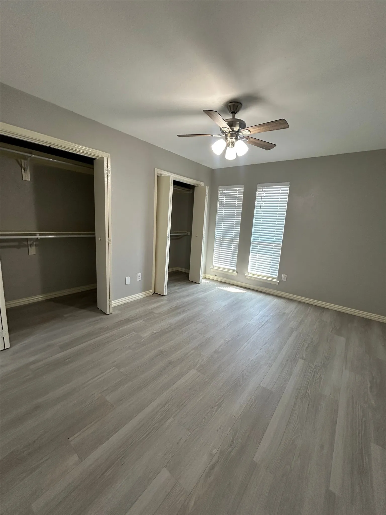 Grey wood-finish flooring, two large windows with blinds, two open closet spaces, and a ceiling fan with integrated lighting