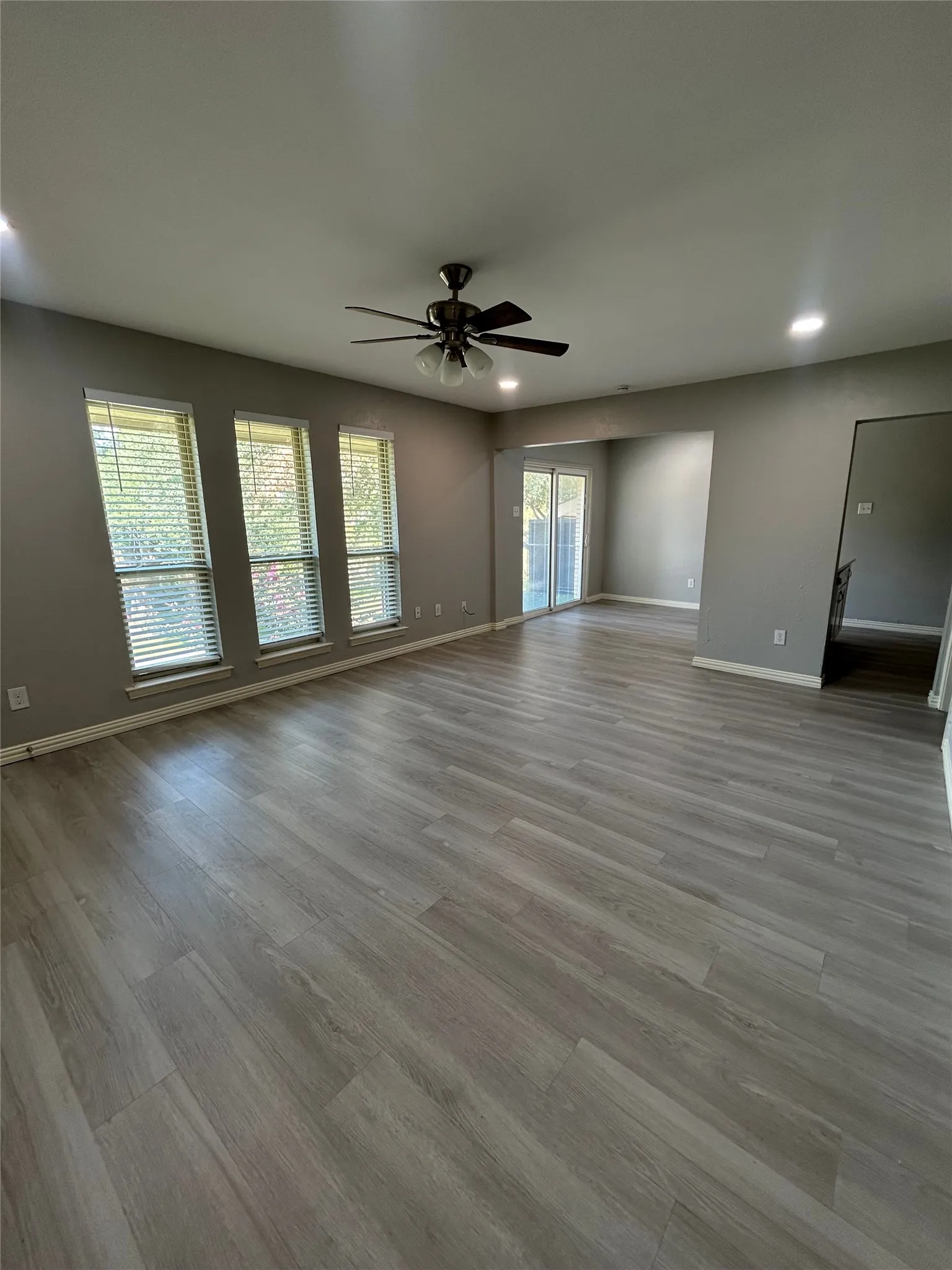Spacious room featuring wood-finish flooring, three large windows with blinds, recessed lighting, and a ceiling fan