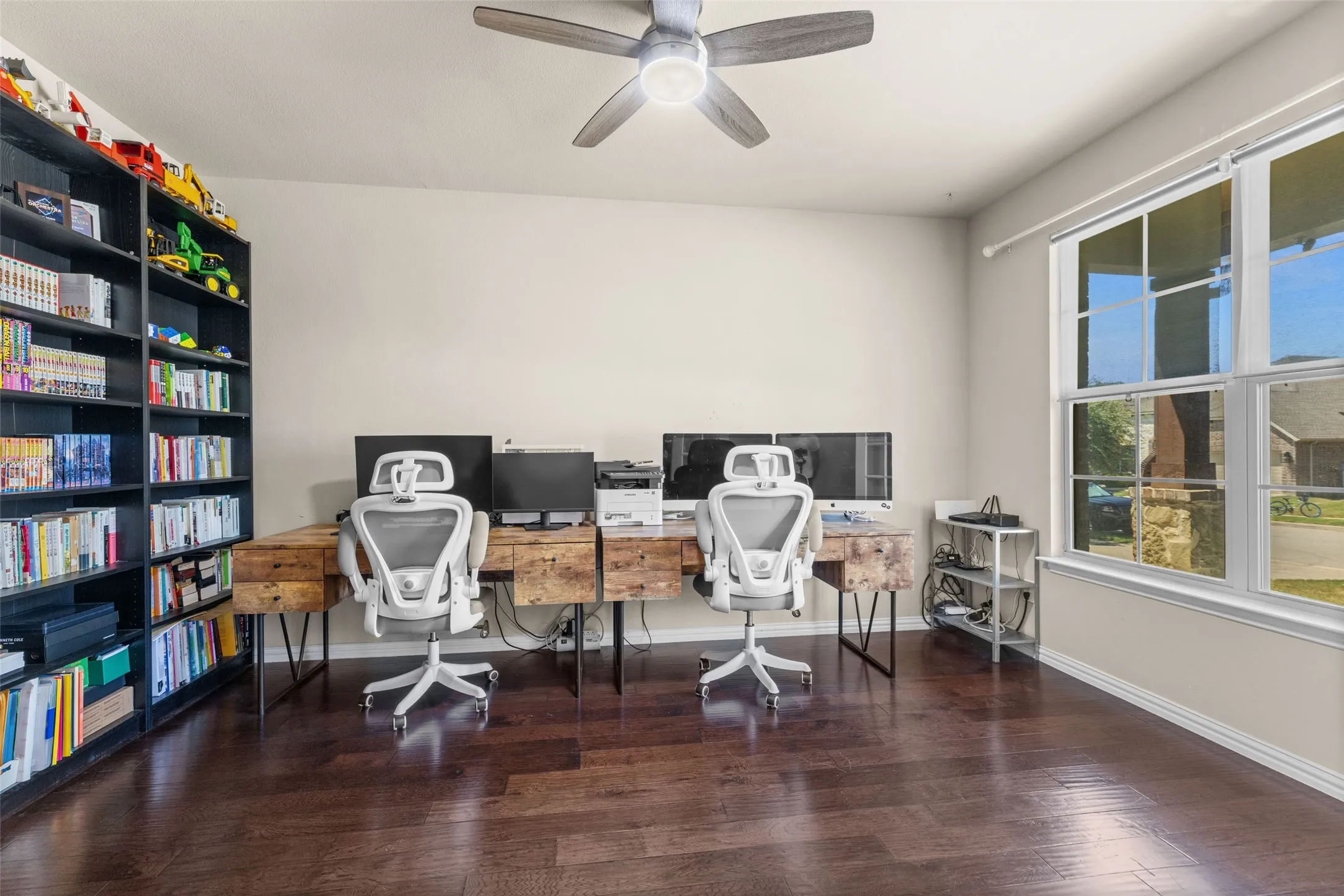 Spacious room featuring wood-finish flooring, a built-in shelving unit, a ceiling fan with integrated lighting, and multiple windows