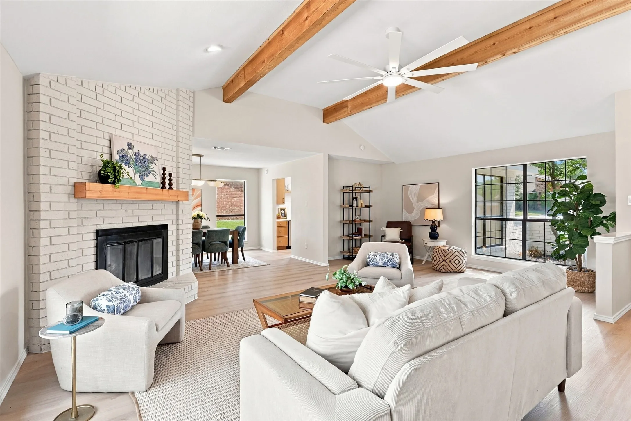 Vaulted ceiling with exposed wood beams, light wood-finish flooring, white brick fireplace, black-framed windows, and an open-concept layout