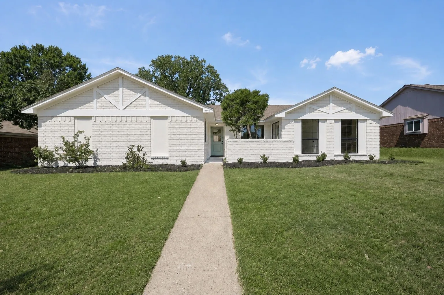 White brick facade with gabled roof lines, featuring black-framed windows, a concrete walkway, and landscaped front yard with shrubs