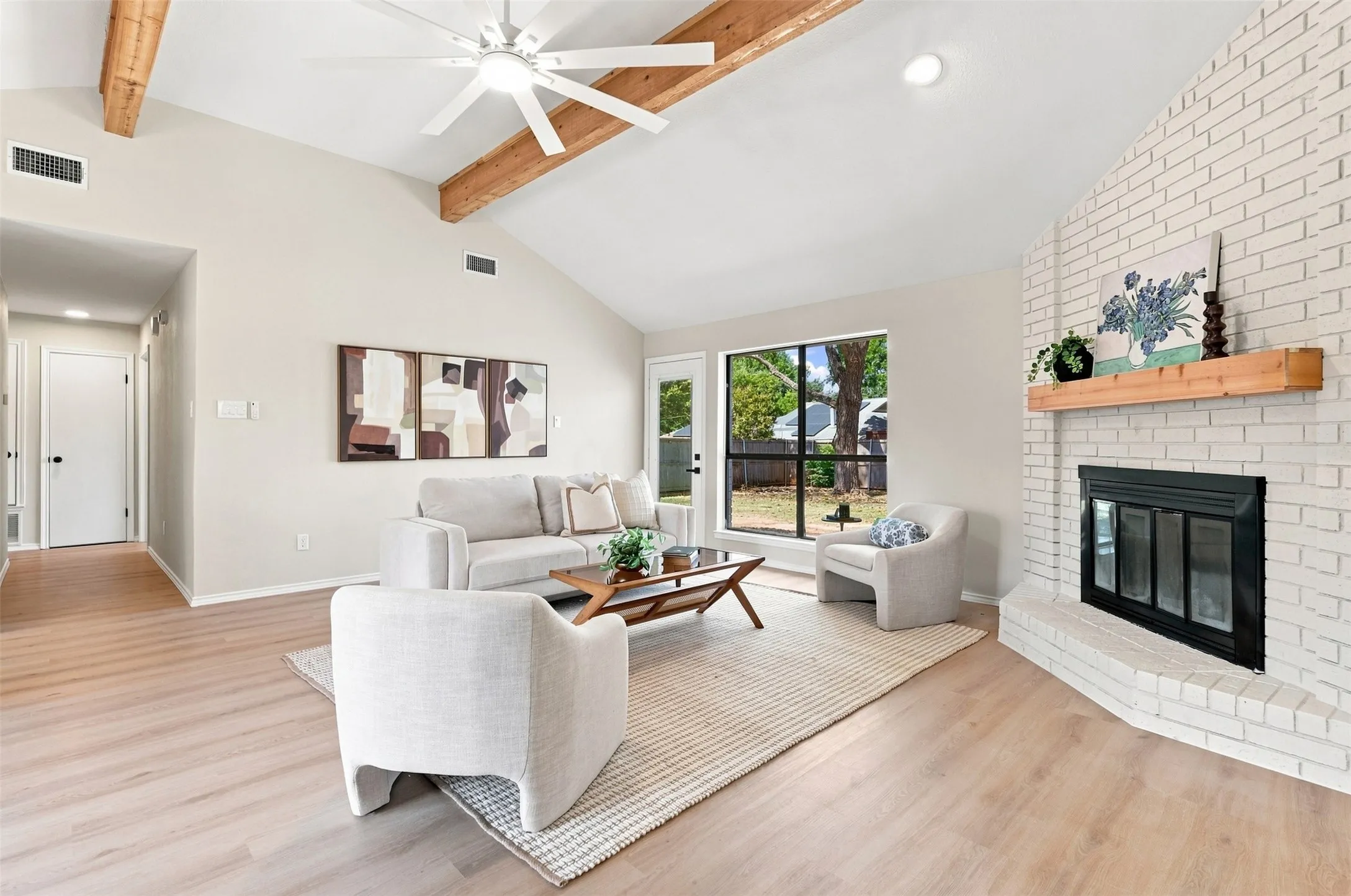 Vaulted ceiling with exposed wood beams and ceiling fan, wood-finish flooring, white brick fireplace with wood mantel, and large window with exterior access