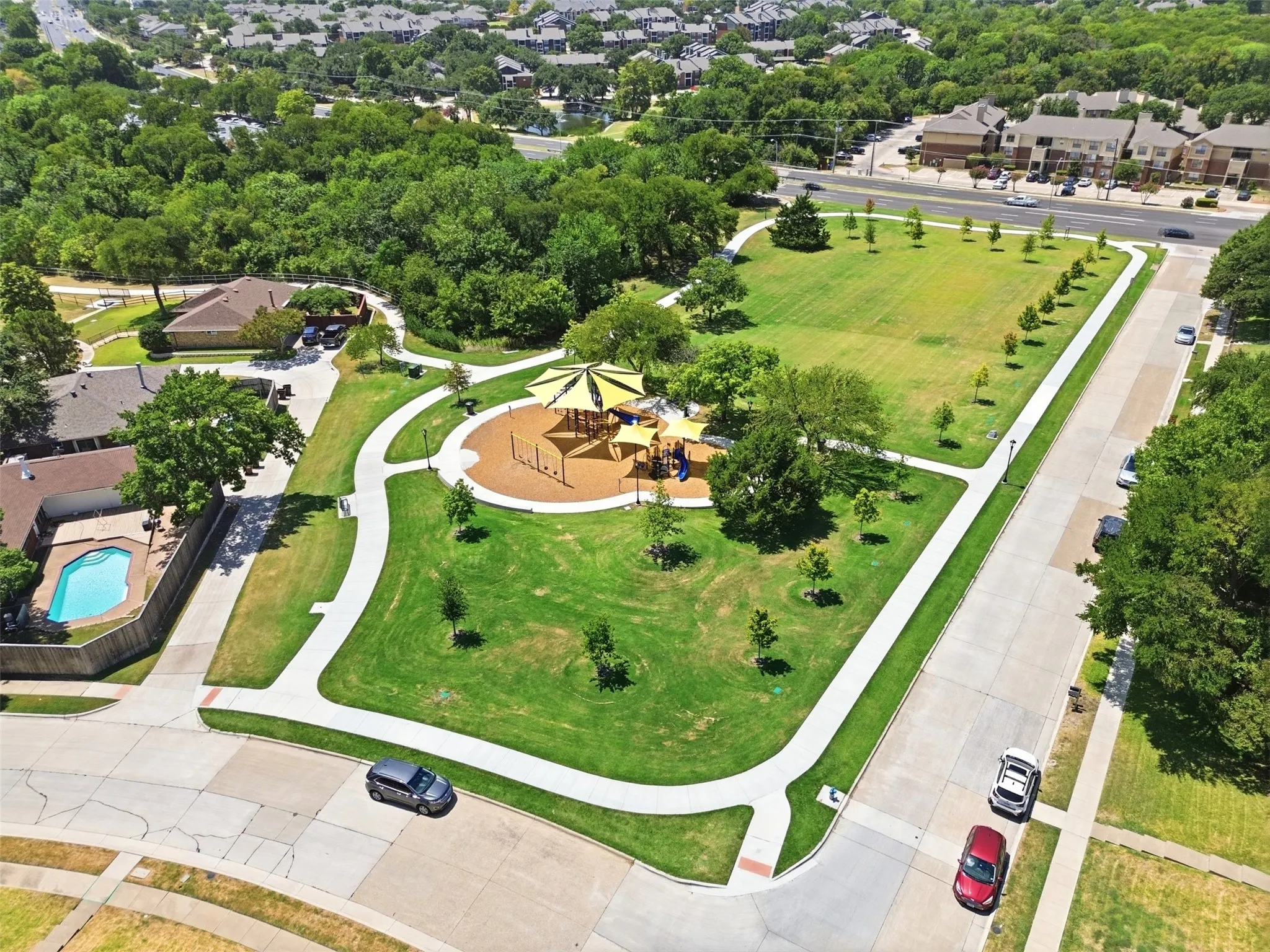 Expansive community park featuring a playground area, green space, and paved walking paths, surrounded by mature trees and residential properties