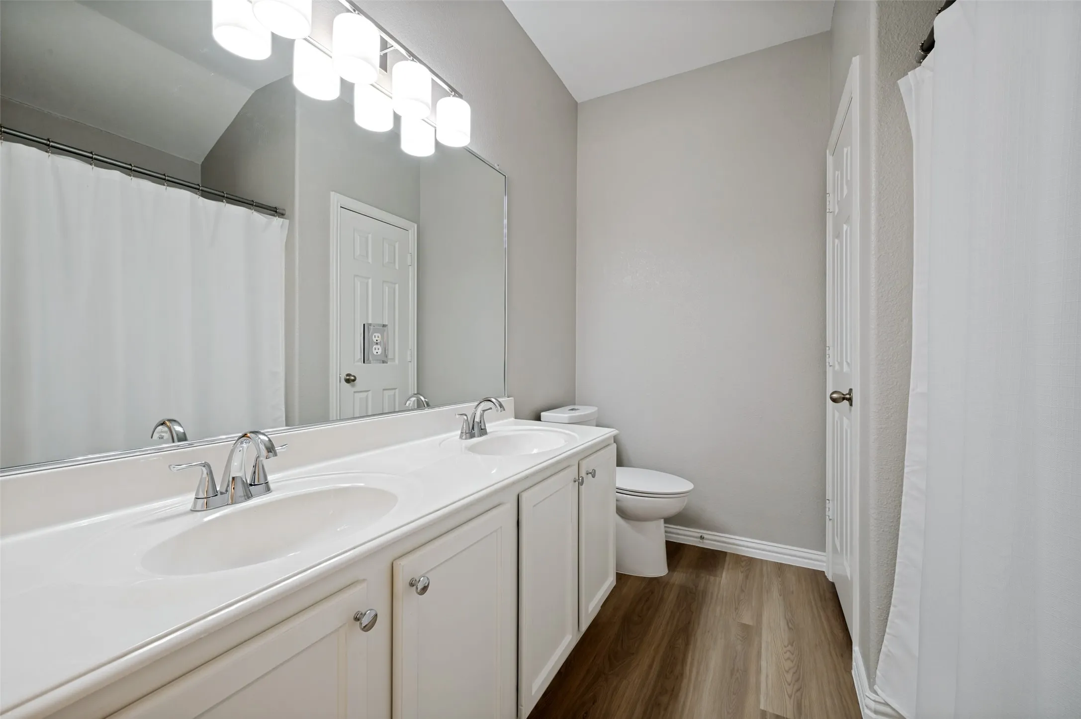 Double vanity with chrome fixtures and integrated sinks, wood-finish flooring, and a large wall-mounted mirror