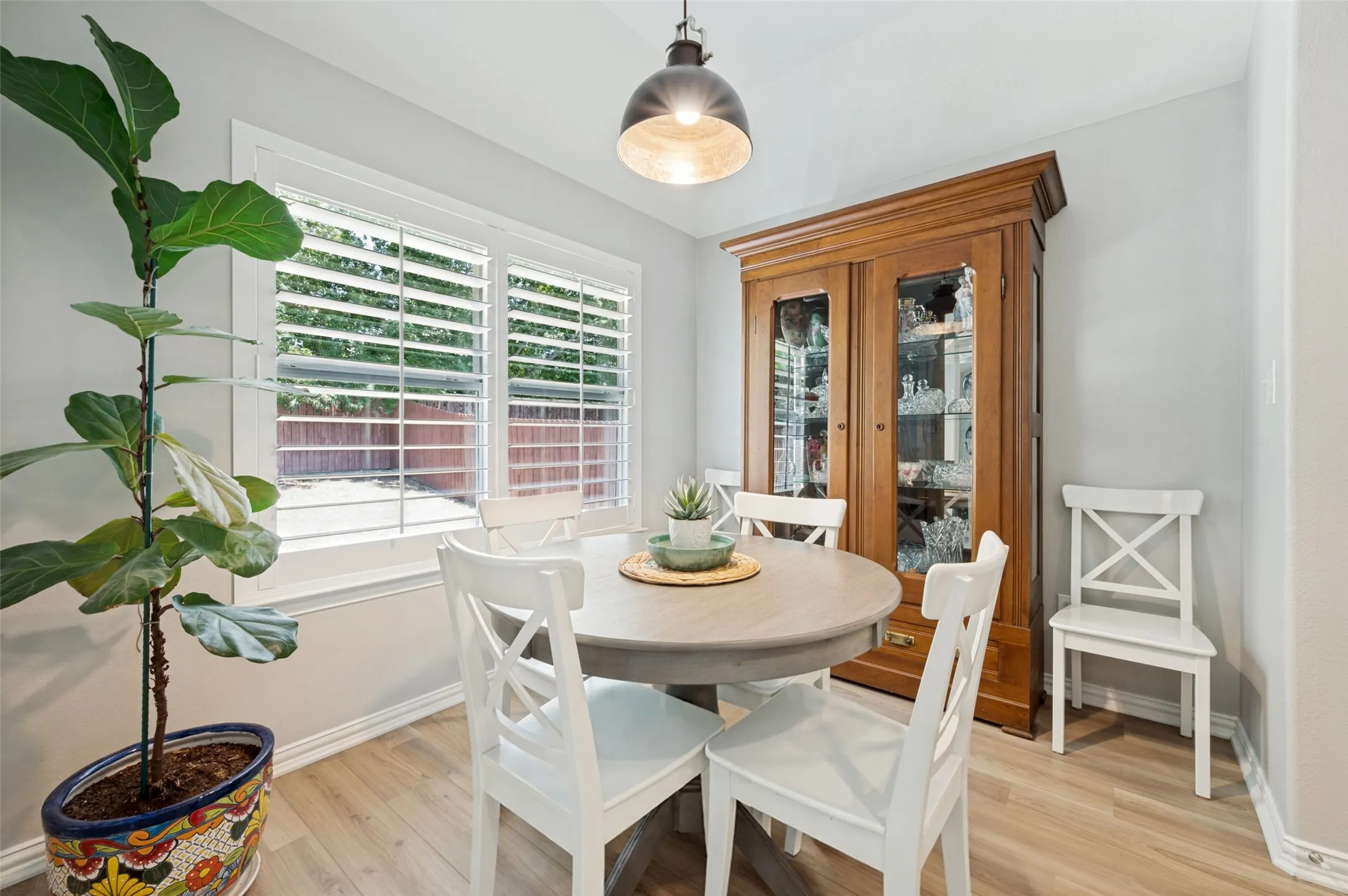 Wood-finish flooring with light-toned walls, featuring a window with white louvered shutters, and a dark-toned suspended light fixture