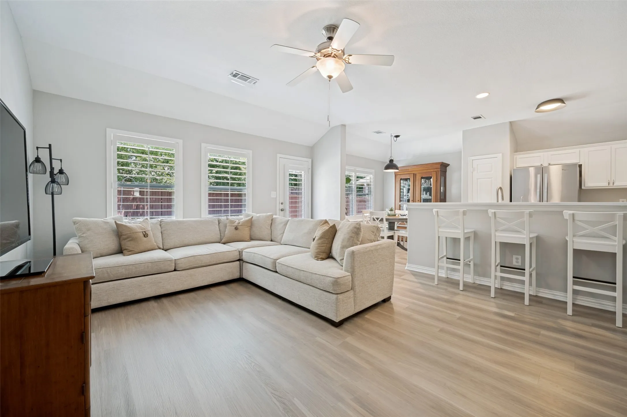 Open-concept living and kitchen area featuring a cream upholstered sectional sofa, wood-finish flooring throughout, stainless steel appliances, white cabinetry, and abundant natural light from multiple windows with plantation shutters