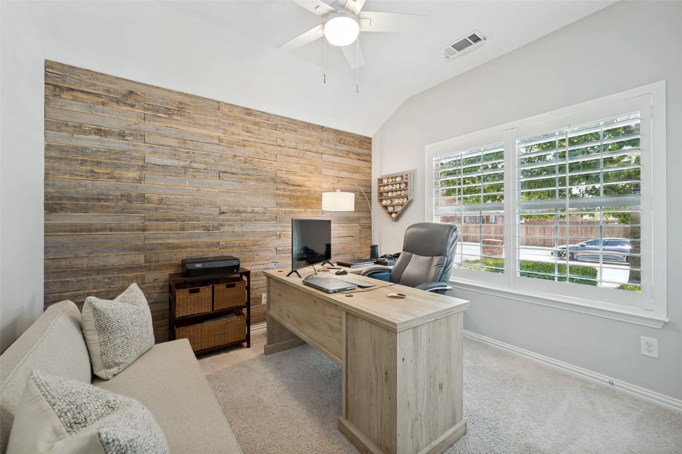 Guest bedroom with a ceiling fan, accent wall with wood paneling, large windows with plantation shutters, and carpeted flooring