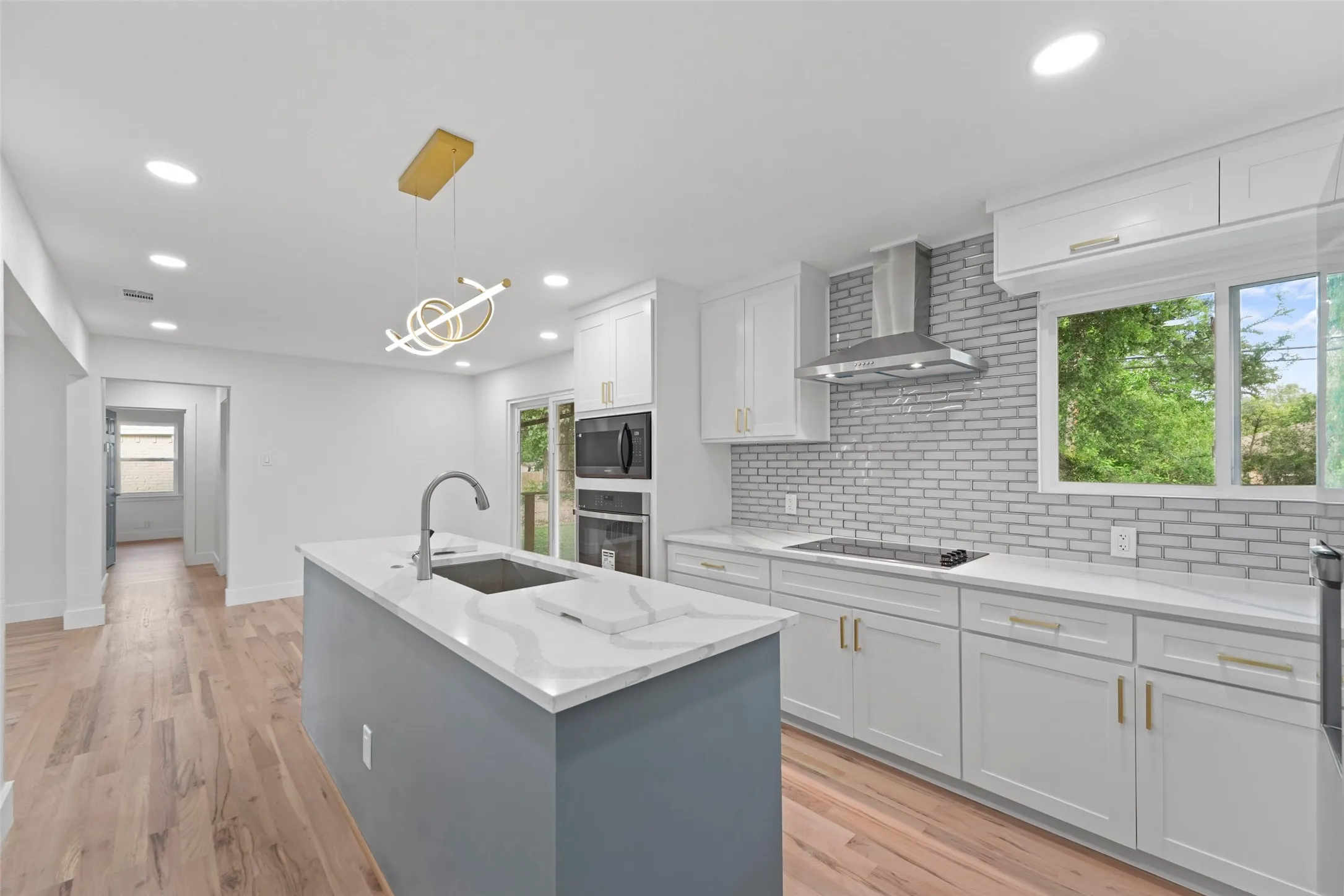The kitchen features light-colored hardwood flooring, a central island with a sink and a modern gold-toned light fixture above, and white cabinetry with a light-colored countertop