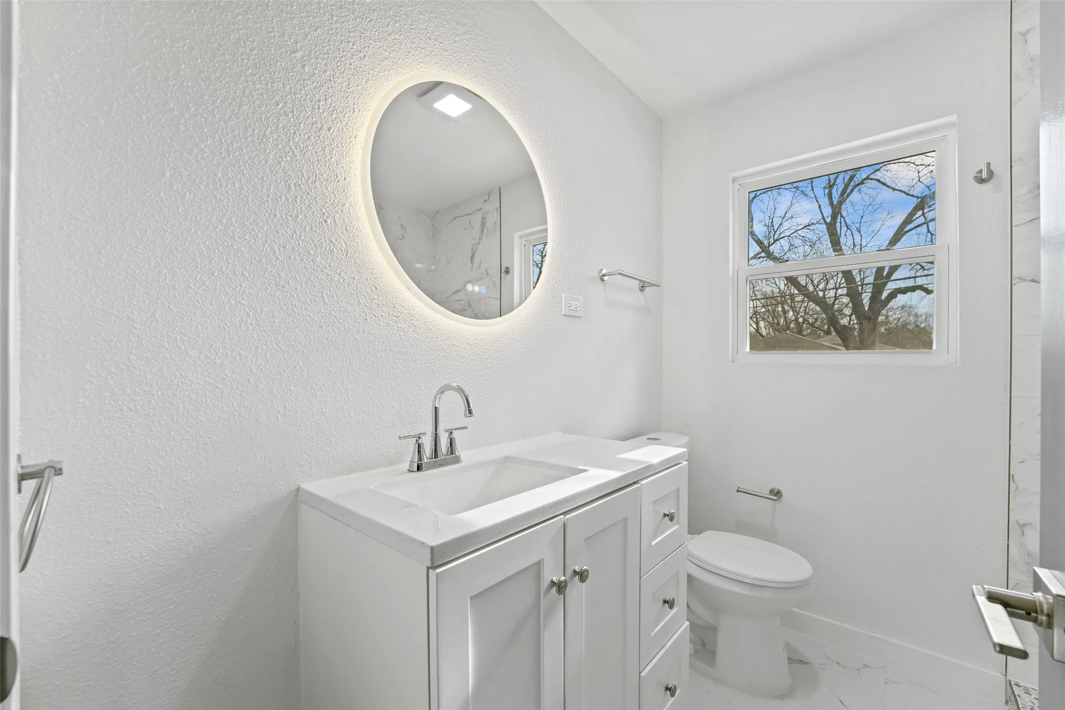 The bathroom features a modern vanity with a white countertop and chrome faucet, an illuminated oval mirror, and a window providing natural light