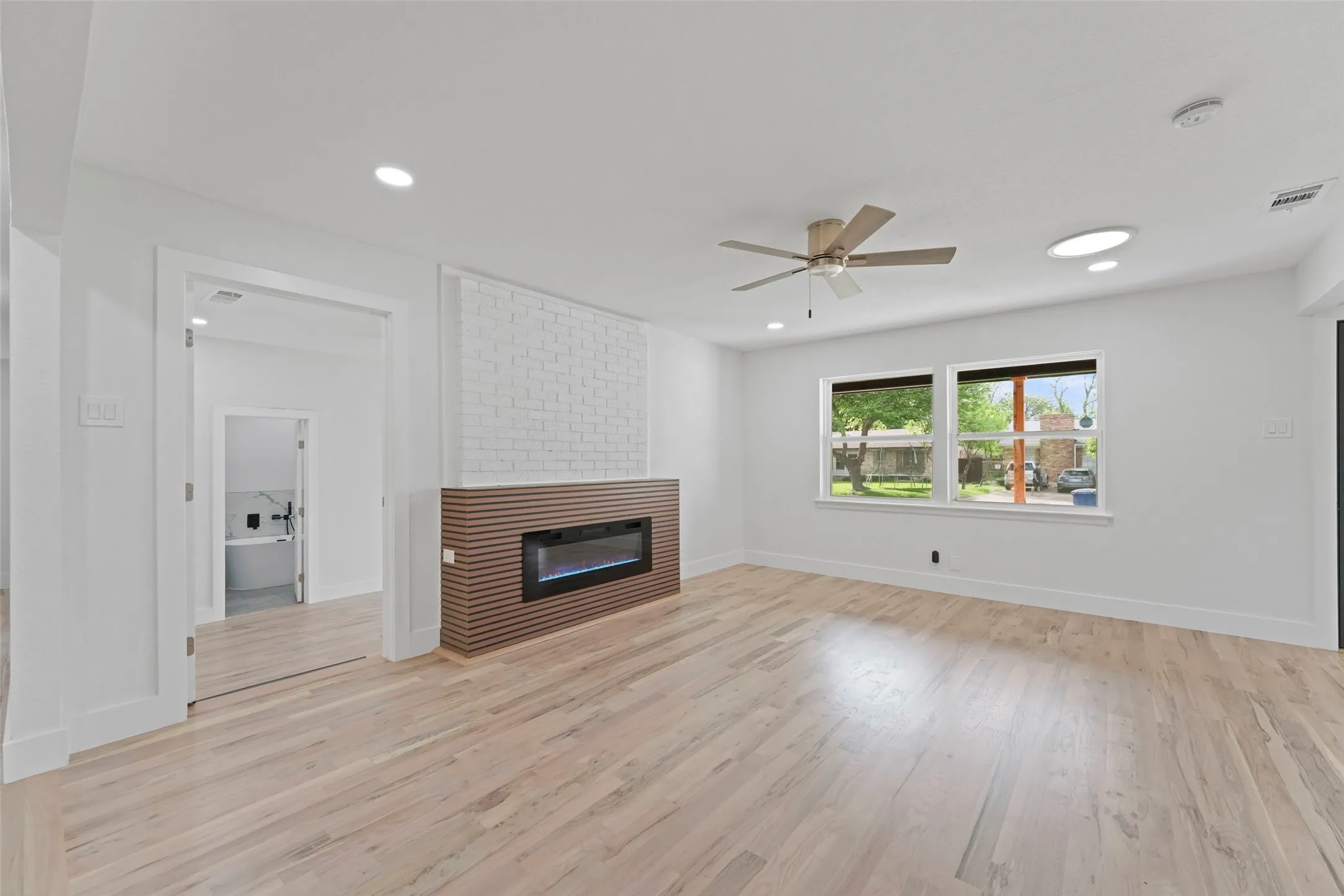 This inviting room features light-toned hardwood flooring, a contemporary fireplace with a slatted wood surround and white brick accent, and a ceiling fan