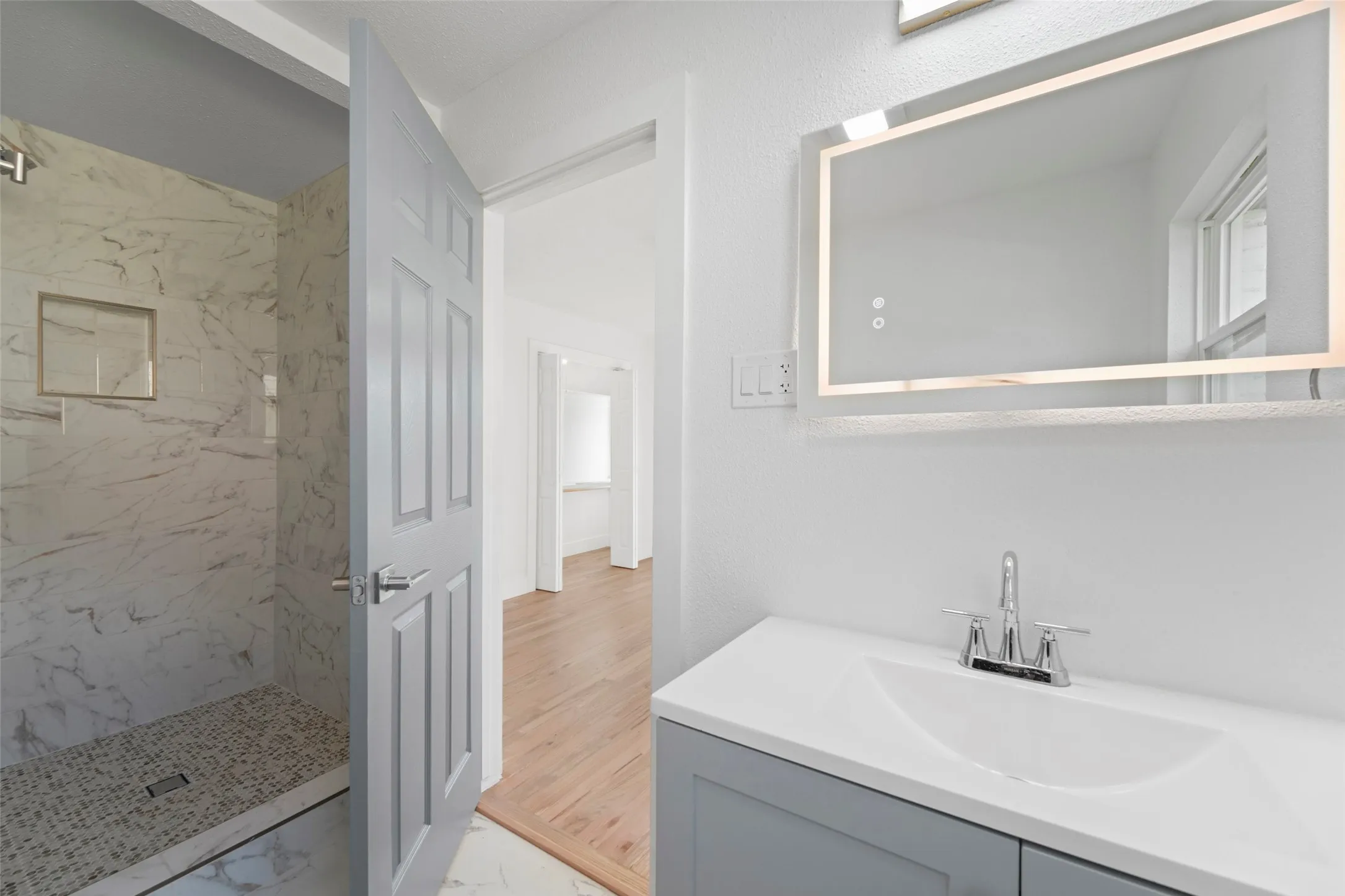 The bathroom features a modern vanity with a white countertop and a lighted mirror, alongside a shower with marble-patterned tiling and a tiled floor
