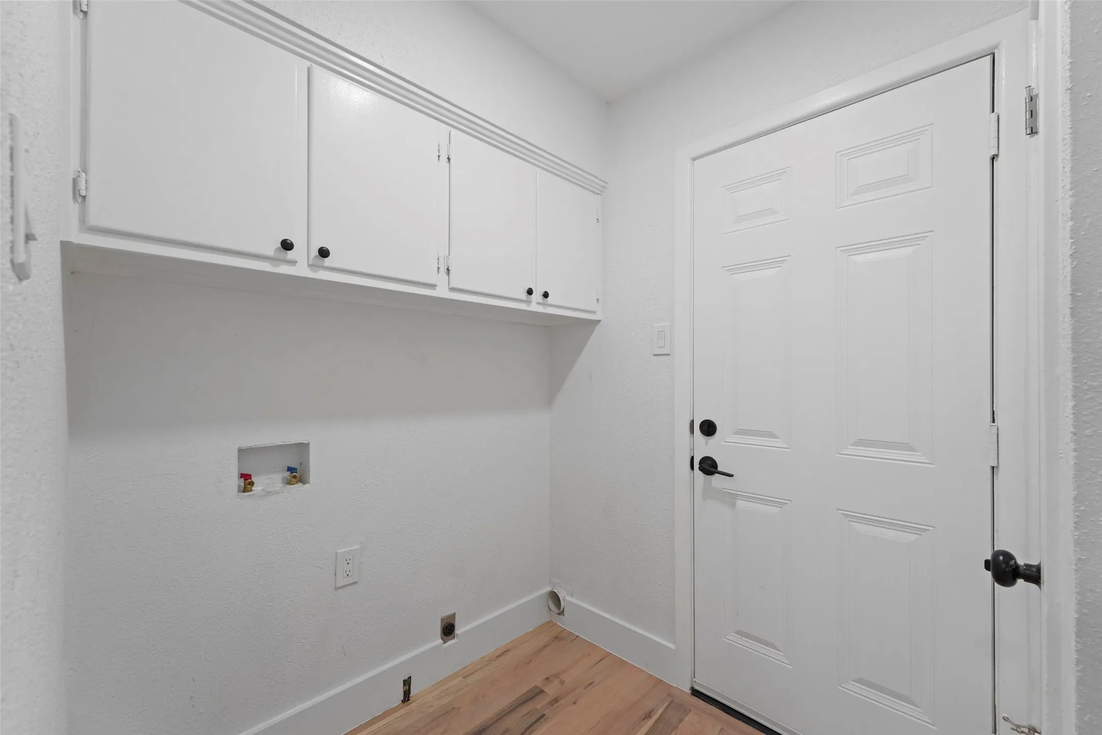 Utility room with white shaker style cabinets, a white paneled door with a black handle, and light wood flooring