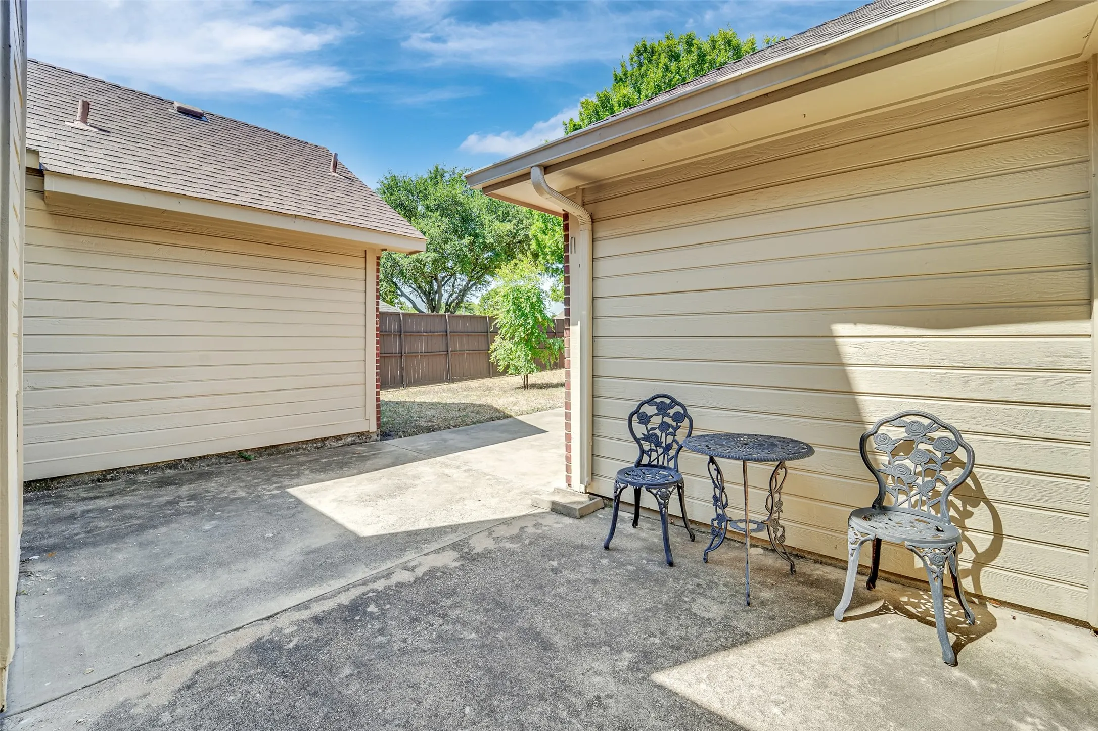 Covered carport with concrete flooring and open-air seating area, framed by horizontal siding and mature landscaping
