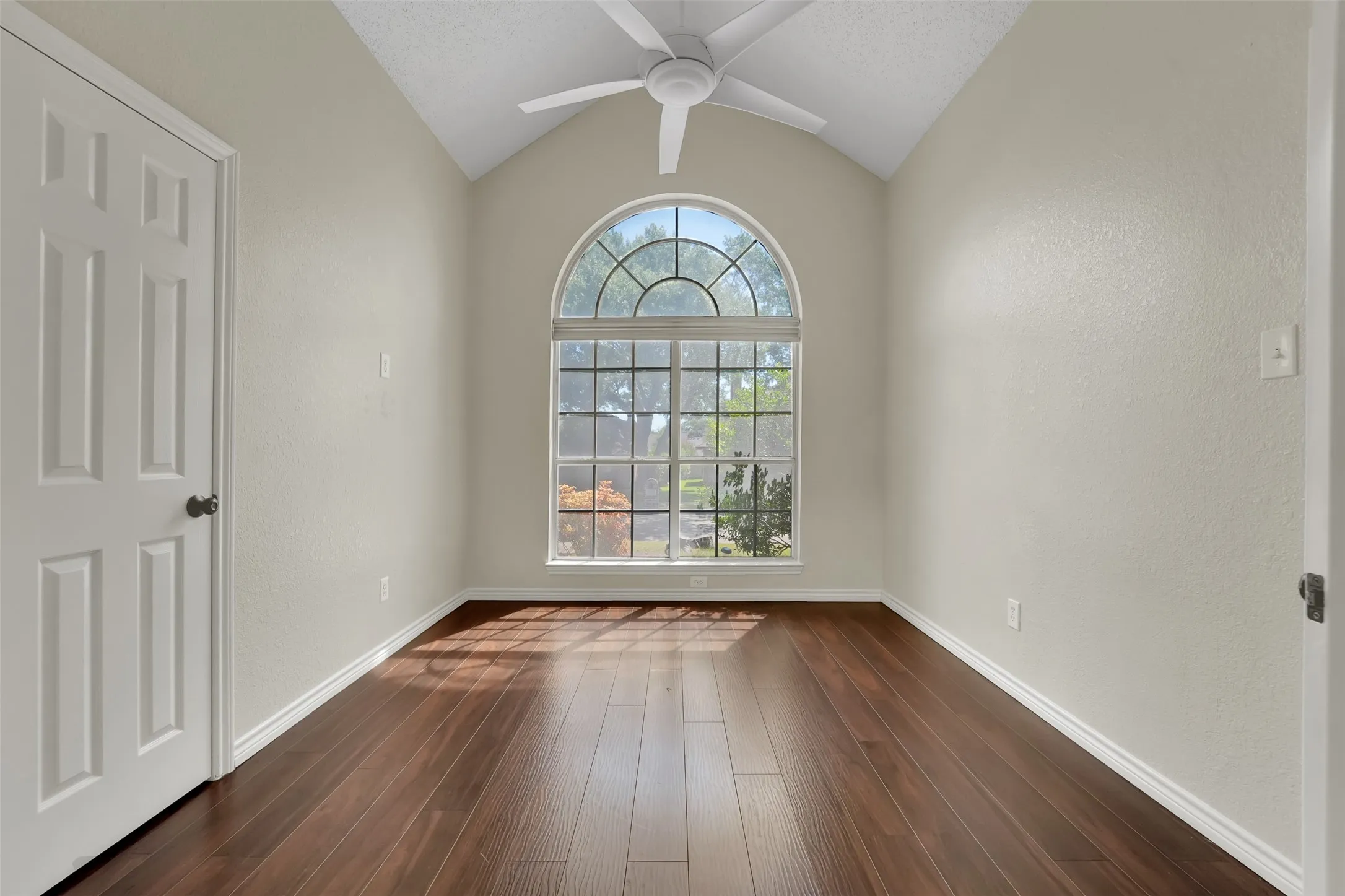 Bright, light-filled room featuring soaring vaulted ceilings with ceiling fan, rich wood-finish flooring throughout, and an exceptional arched transom window with multiple panes flooding the space with natural light