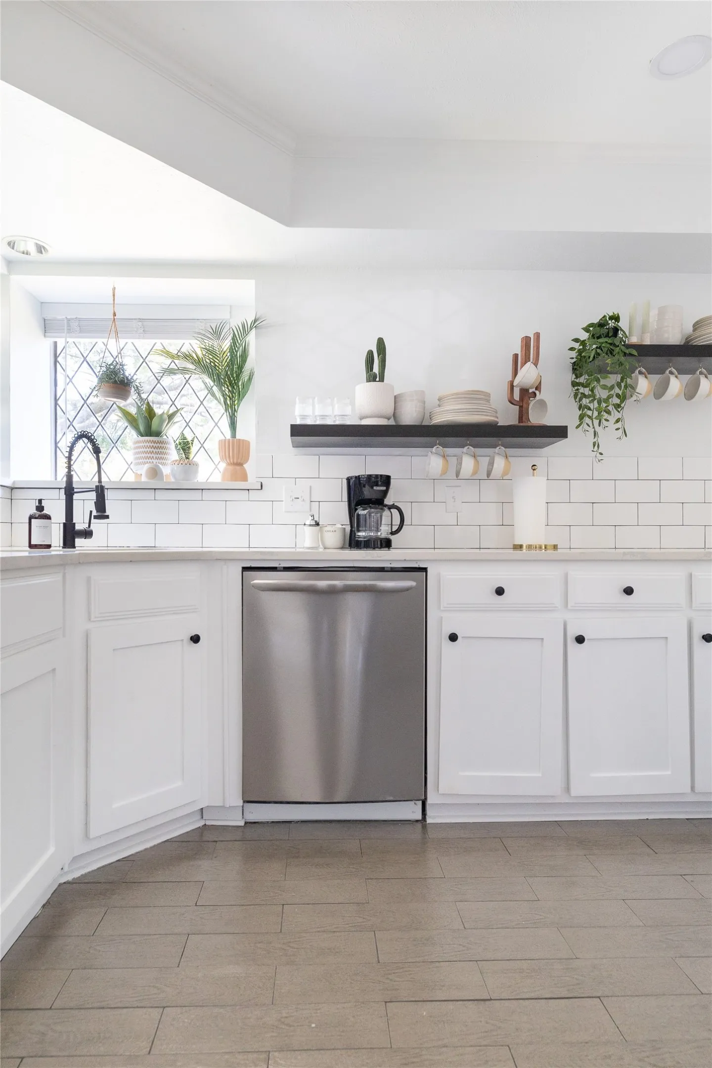 White shaker-style cabinetry with black knob hardware, stainless steel dishwasher, white subway tile backsplash, white countertops, and wood-finish flooring