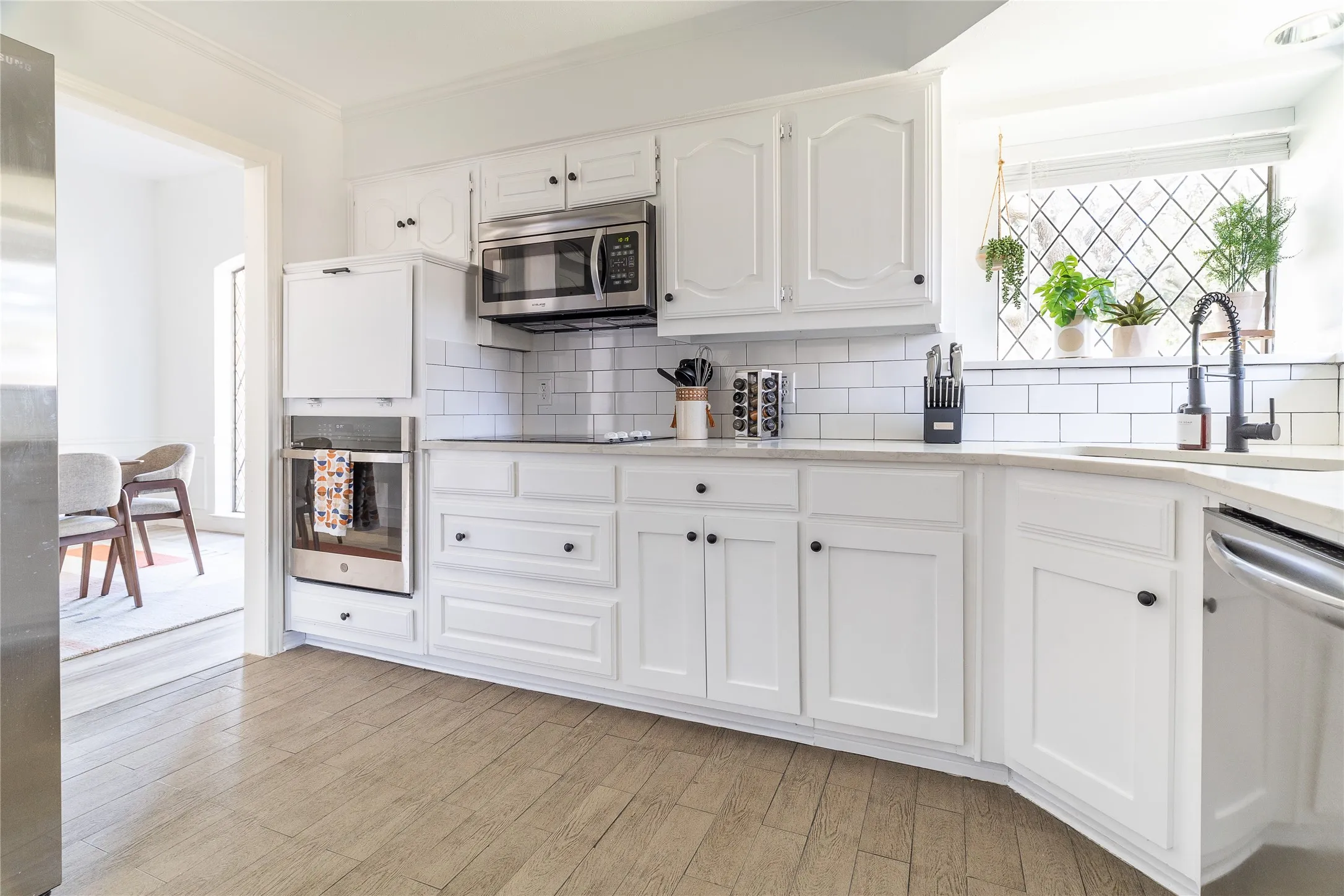 White cabinetry with black hardware, subway tile backsplash, stainless steel microwave, built-in oven, and wood-finish flooring