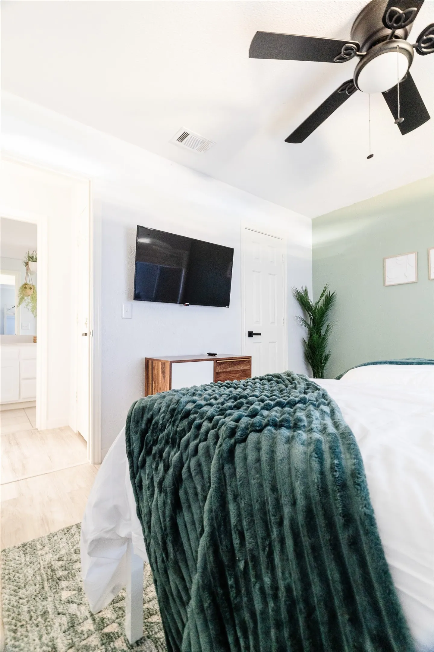 Ceiling fan with integrated lighting, wall-mounted television, wood-finish flooring, light-toned walls, and a door with matte black hardware