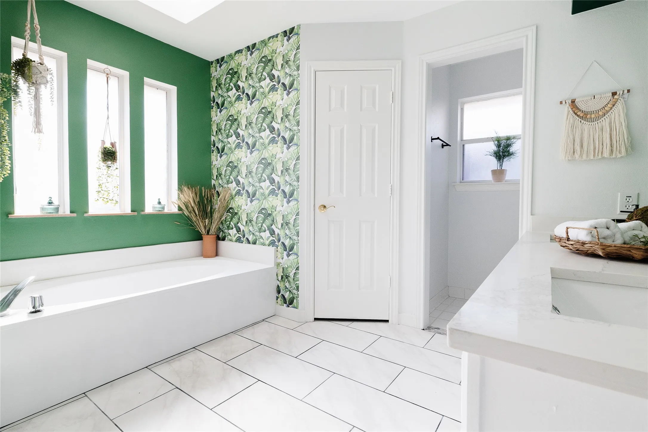 Soaking tub with a polished chrome fixture, white rectangular floor tiles, green accent wall, and a vanity with a white countertop