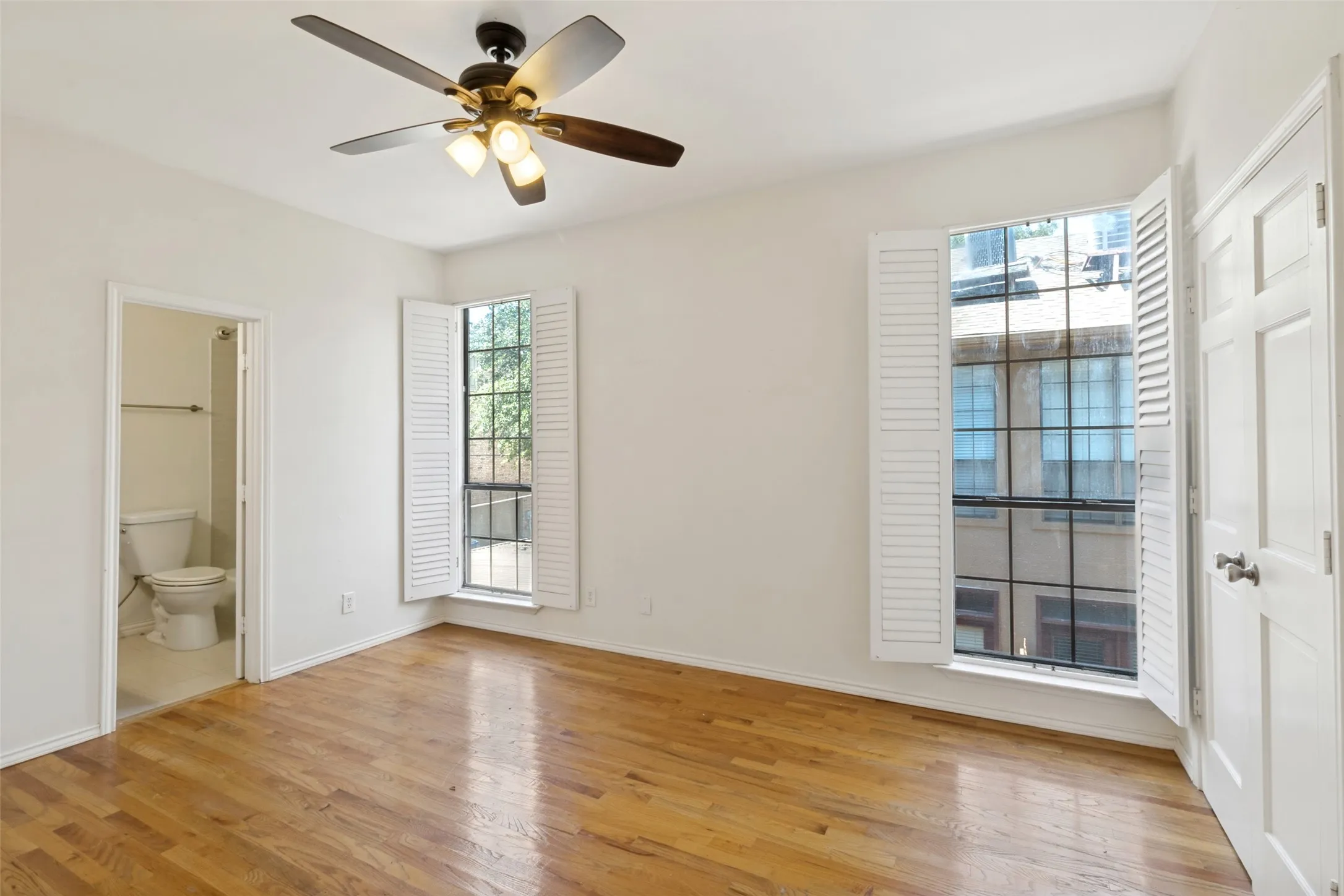 Wood-finish flooring, two windows with louvered shutters, ceiling fan with integrated lighting, white interior doors