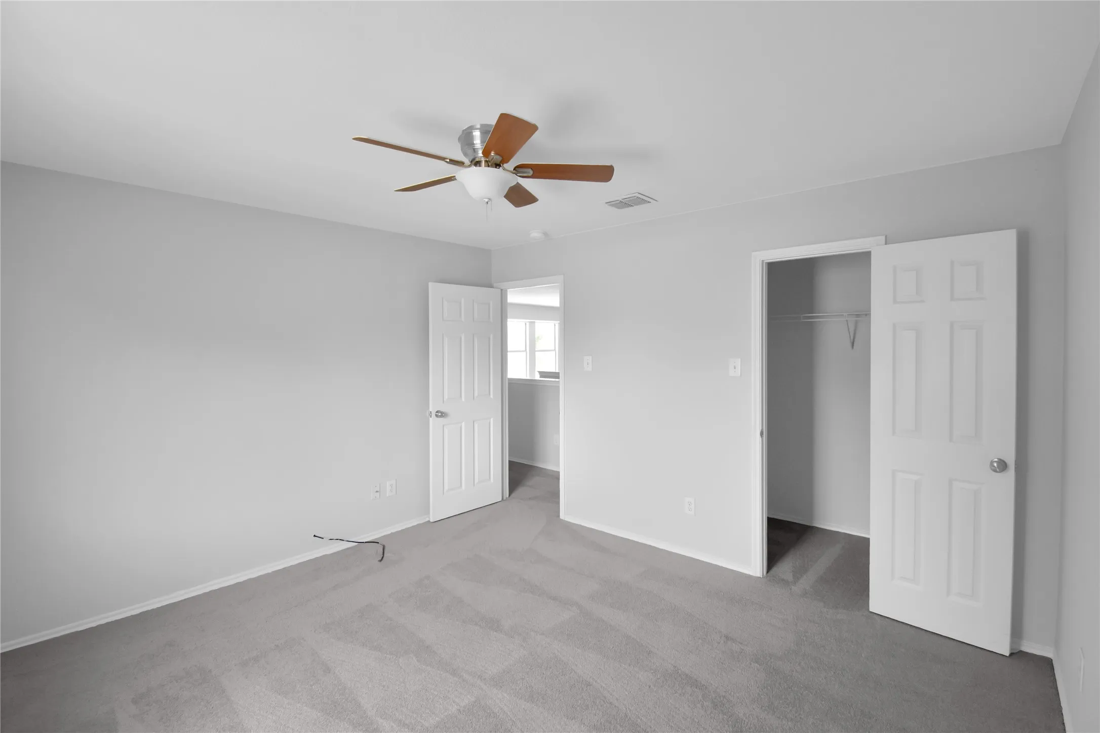 Neutral gray wall finish, gray carpet flooring, ceiling fan with wood-finish blades, white six-panel interior doors, and a built-in closet with shelving