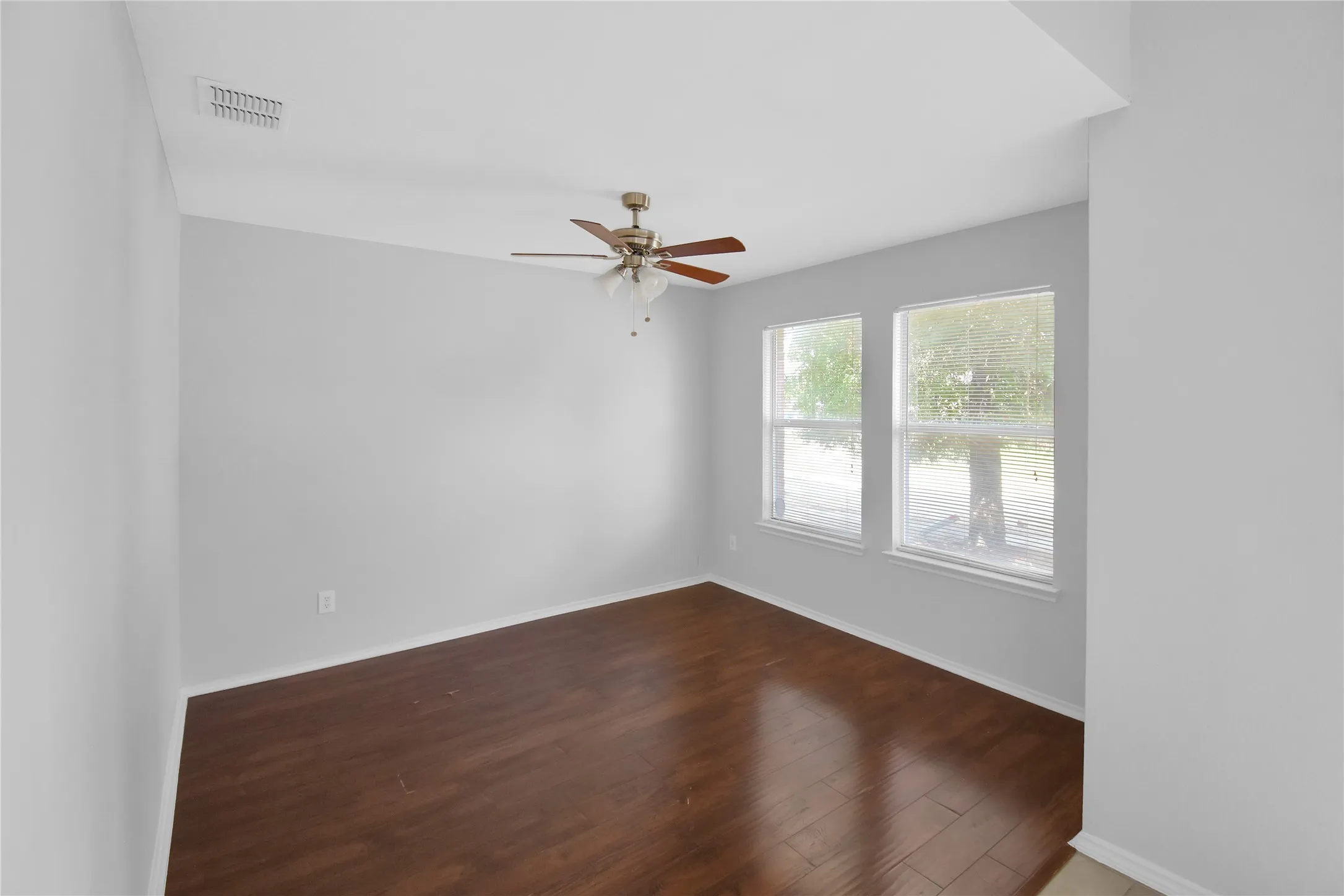 Bright corner bedroom showcasing rich wood-finish flooring, abundant natural light from dual windows, ceiling fan with integrated lighting, and pristine white walls and ceiling