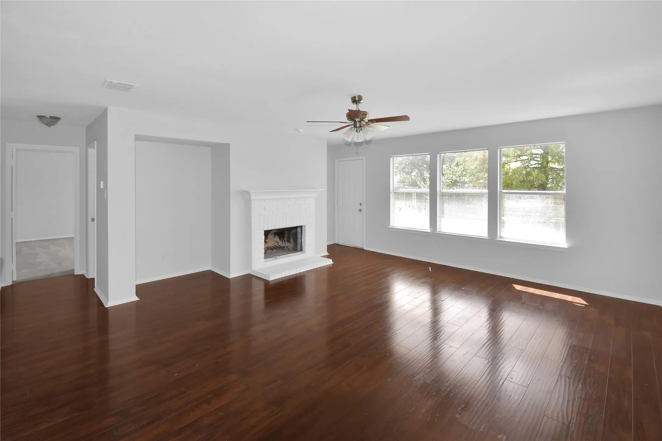 Bright, open living area showcasing rich wood-finish flooring throughout, a ceiling fan with brushed metal finish, and abundant natural light from triple glass doors and windows