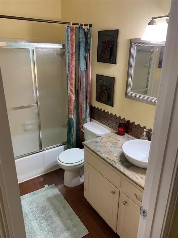 Bathroom featuring a built-in vanity with a stone-finish countertop and vessel sink, wood-finish flooring, a toilet, and a shower-over-tub with a sliding glass enclosure