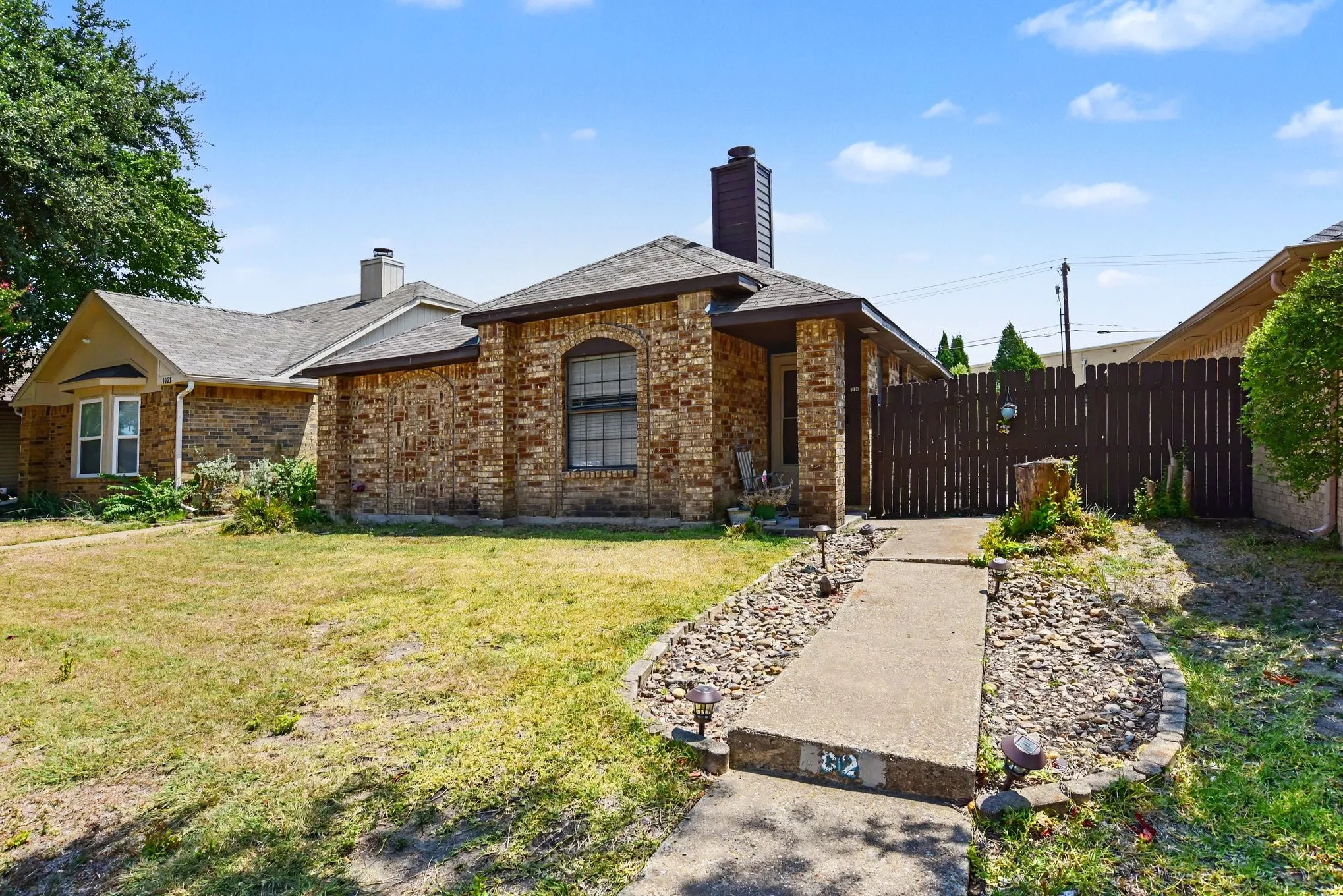 Angled front exterior perspective displaying mature yard trees, brick framing, and paved entrance path.