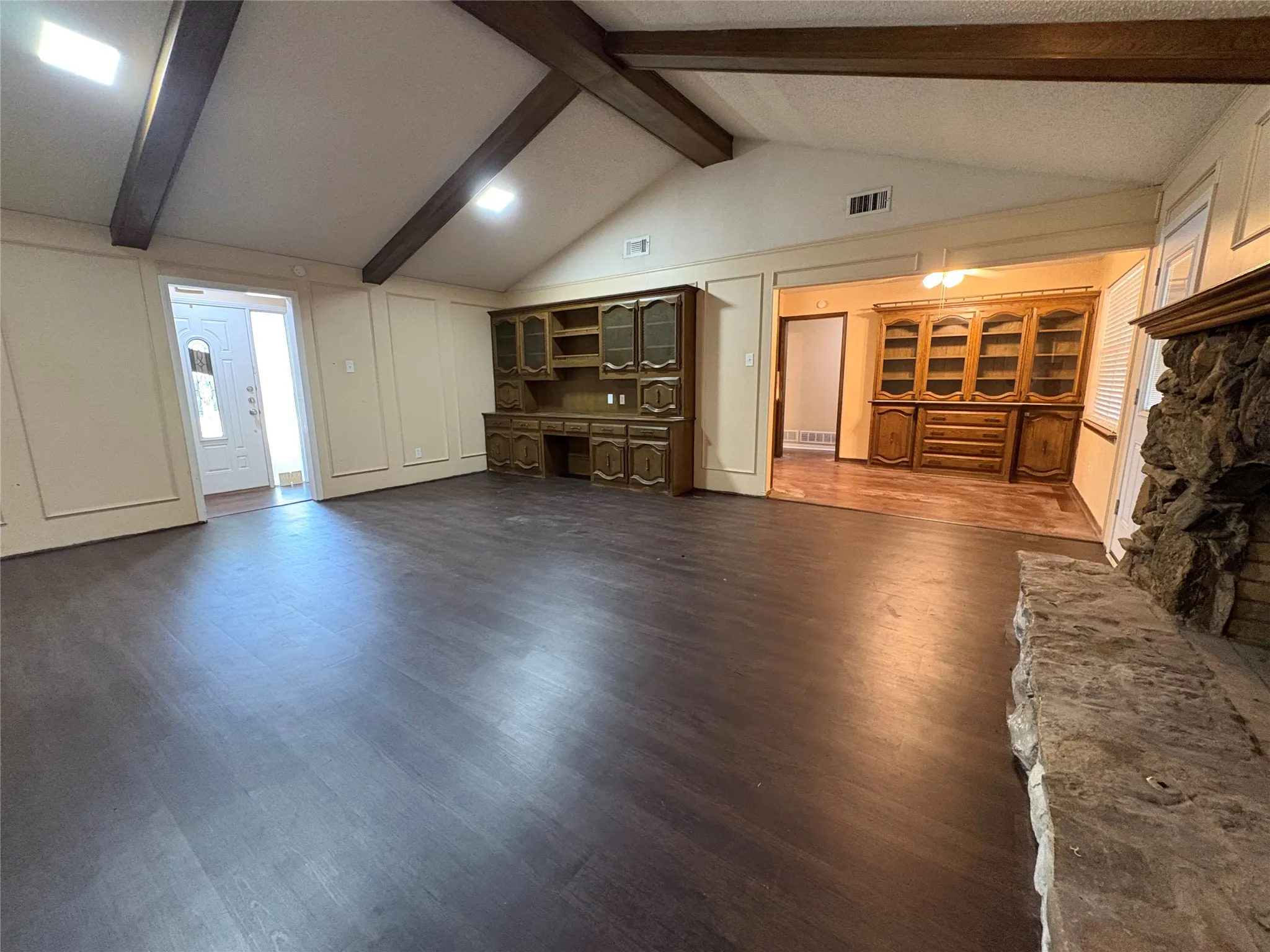 Vaulted ceiling with exposed wood beams, wood-finish flooring, built-in custom cabinetry, and a stone-faced fireplace with a stone hearth