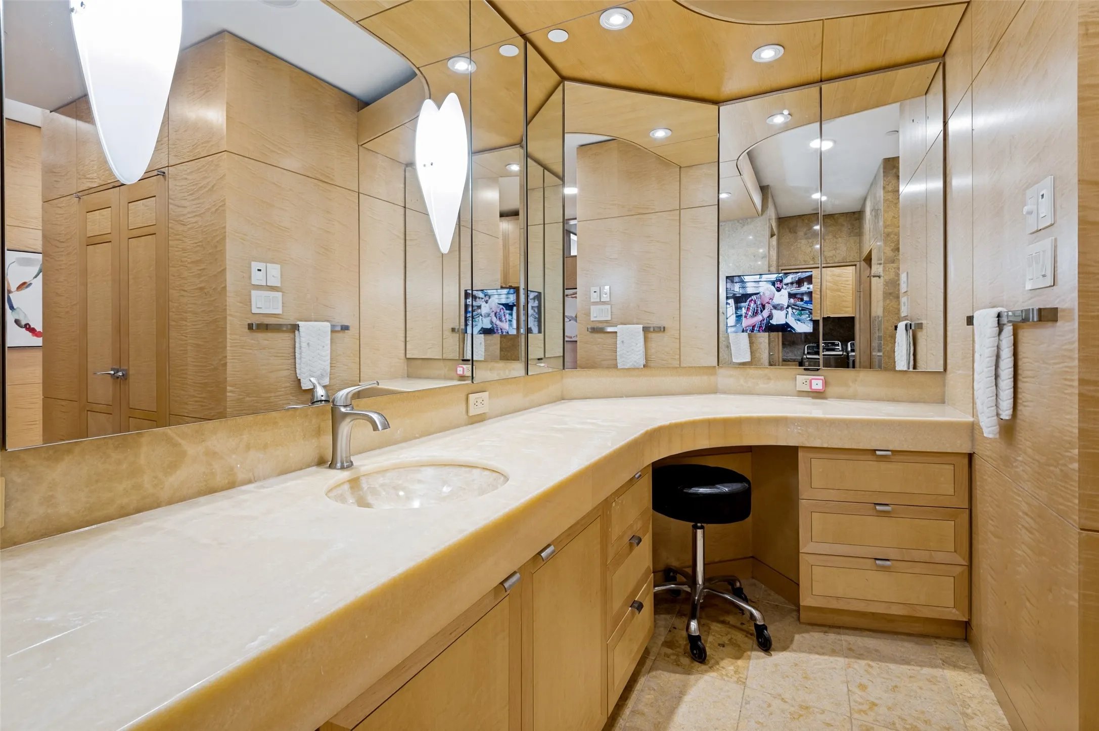 Curved countertop with integrated sink, recessed lighting, wood-finish cabinetry, and vanity mirror with built-in screen