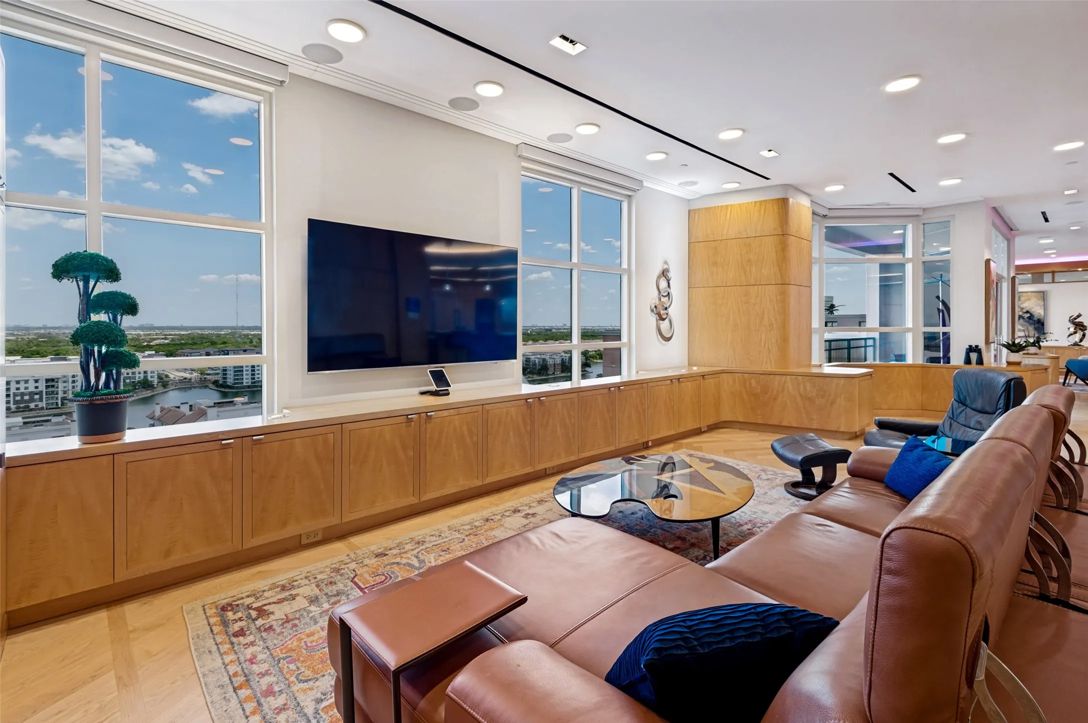 Sophisticated high-rise living room featuring floor-to-ceiling windows with panoramic cityscape vistas, custom wood-finish cabinetry spanning the length of the room, recessed ceiling lighting, wide-plank wood flooring, and a wall-mounted flat-screen television with built-in media infrastructure