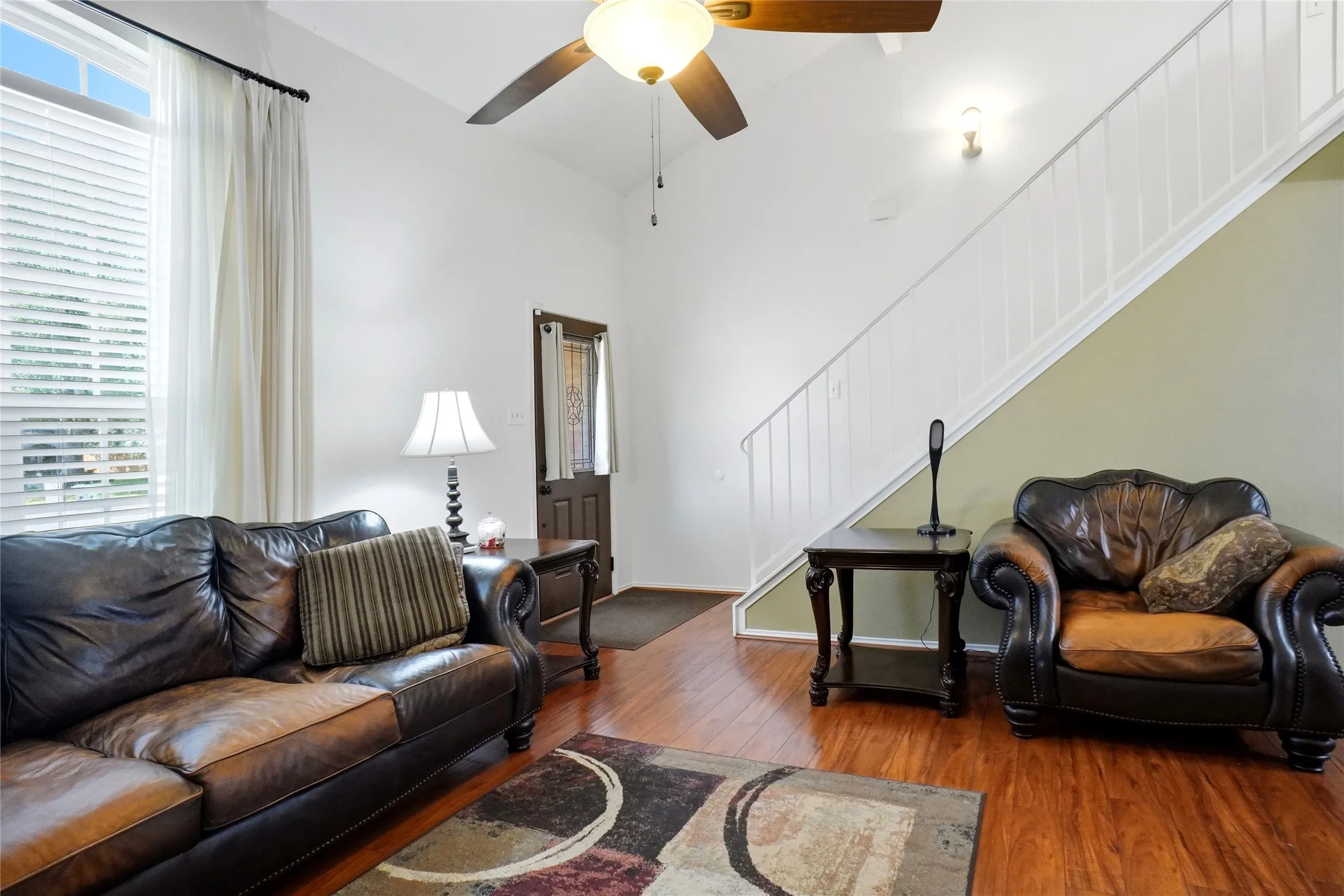 Vaulted ceilings, wood-finish flooring, white window blinds, ceiling fan with integrated lighting, and a white staircase with balusters