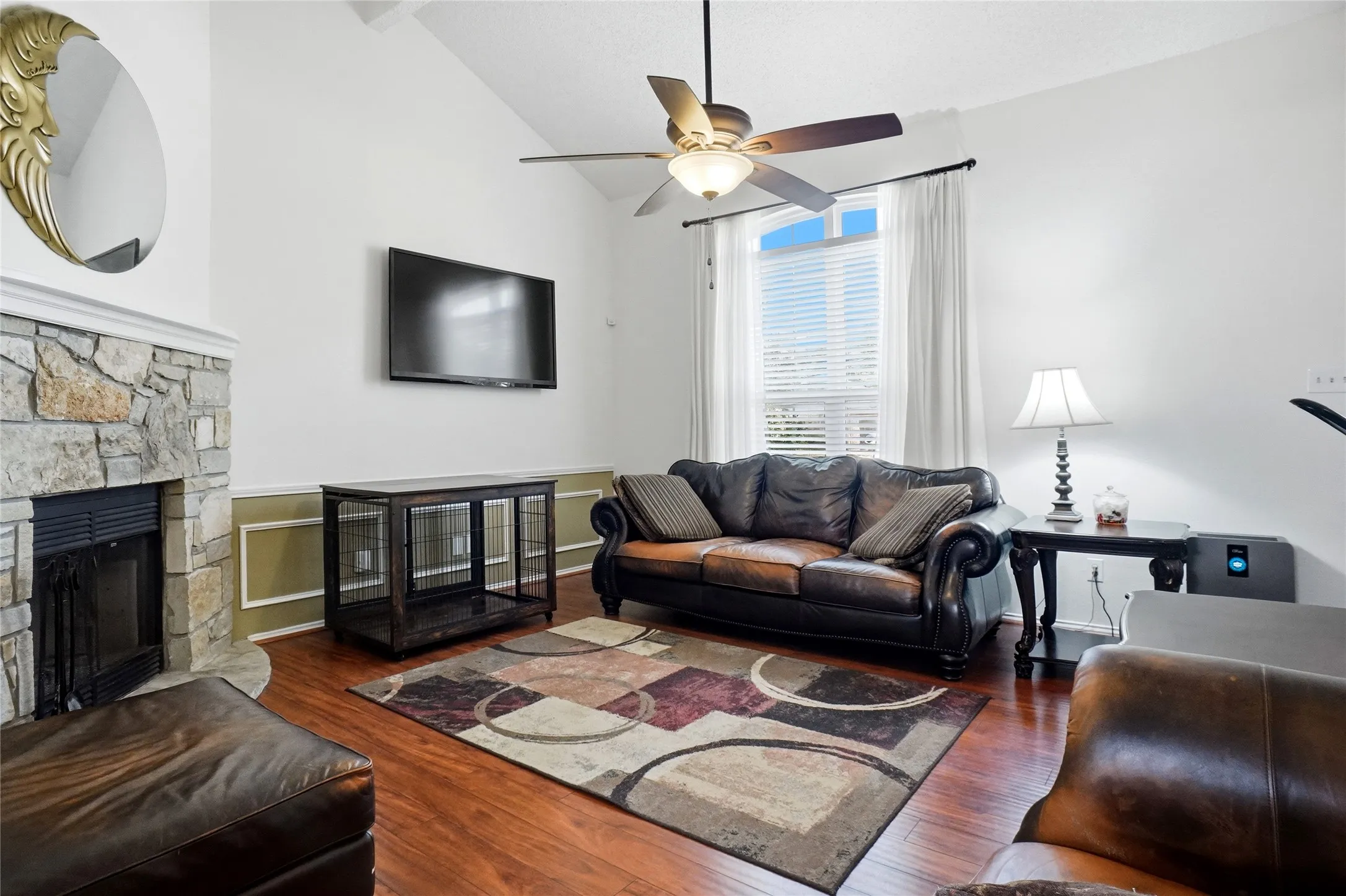 Vaulted ceiling, stone-surround fireplace, wood-finish flooring, ceiling fan, and window with blinds and drapes