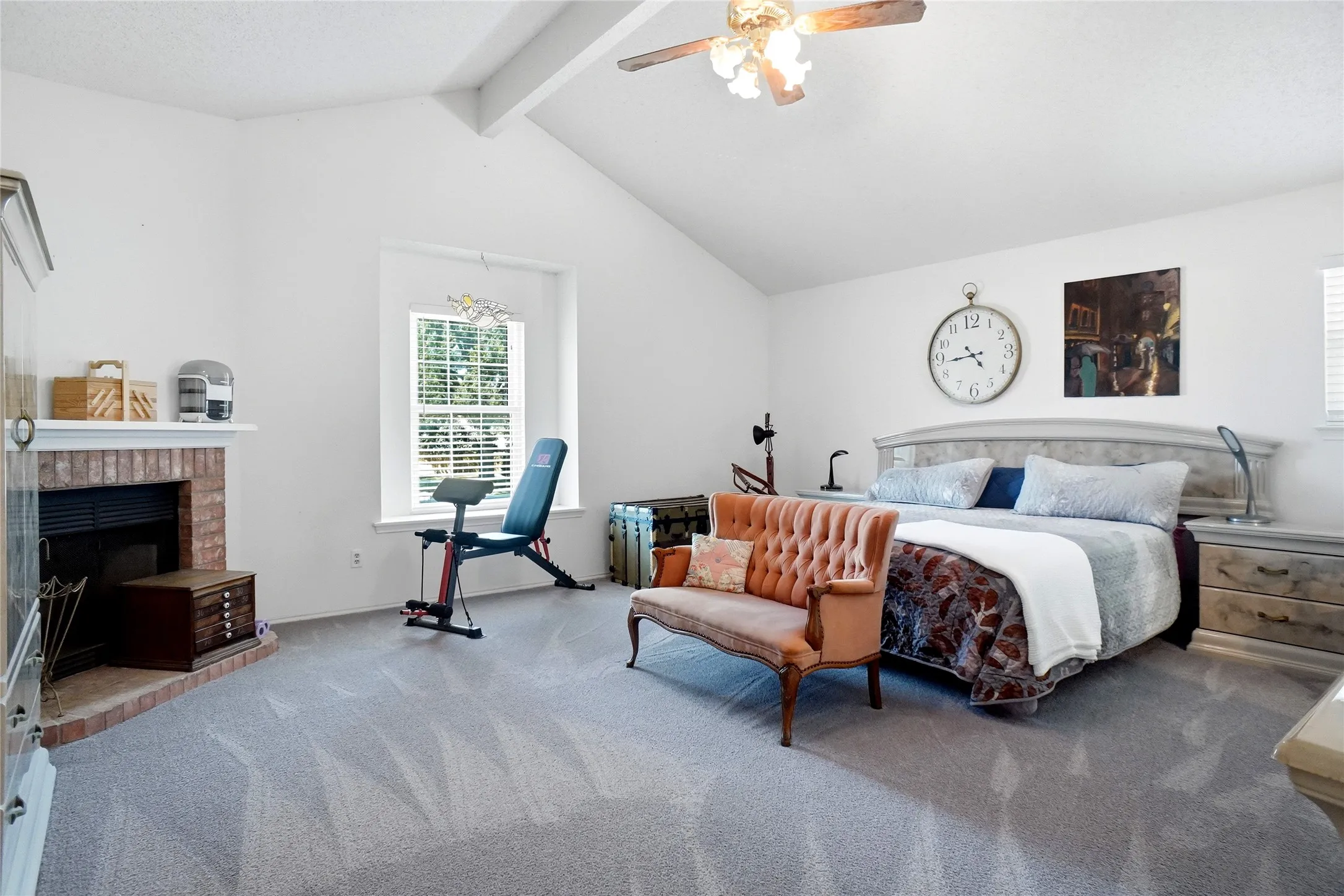 Vaulted white ceiling, brick fireplace, carpeted flooring, and window with outdoor greenery