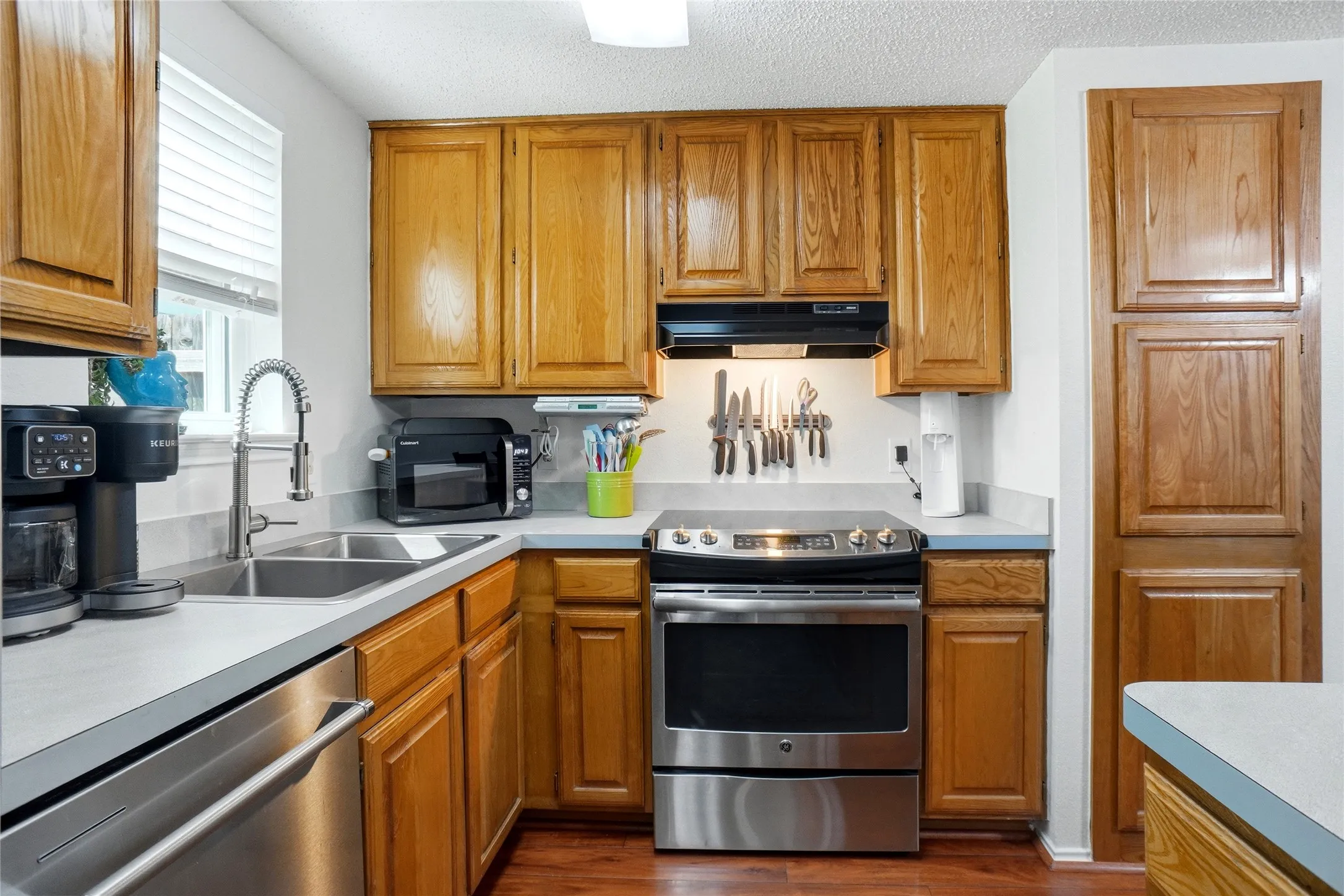 Kitchen featuring wood-finish cabinetry, light-toned countertops, a stainless steel range, a stainless steel dishwasher, and a double basin sink with a gooseneck faucet